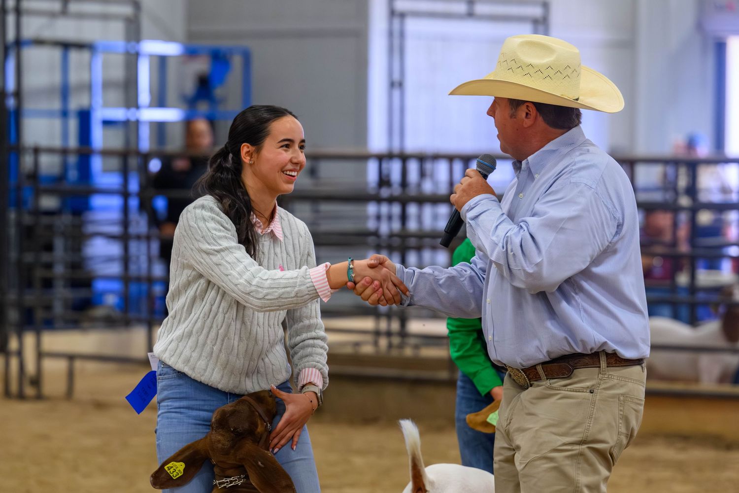 Woman and man in cowboy hat shaking hands at a livestock show, with a brown dog nearby.