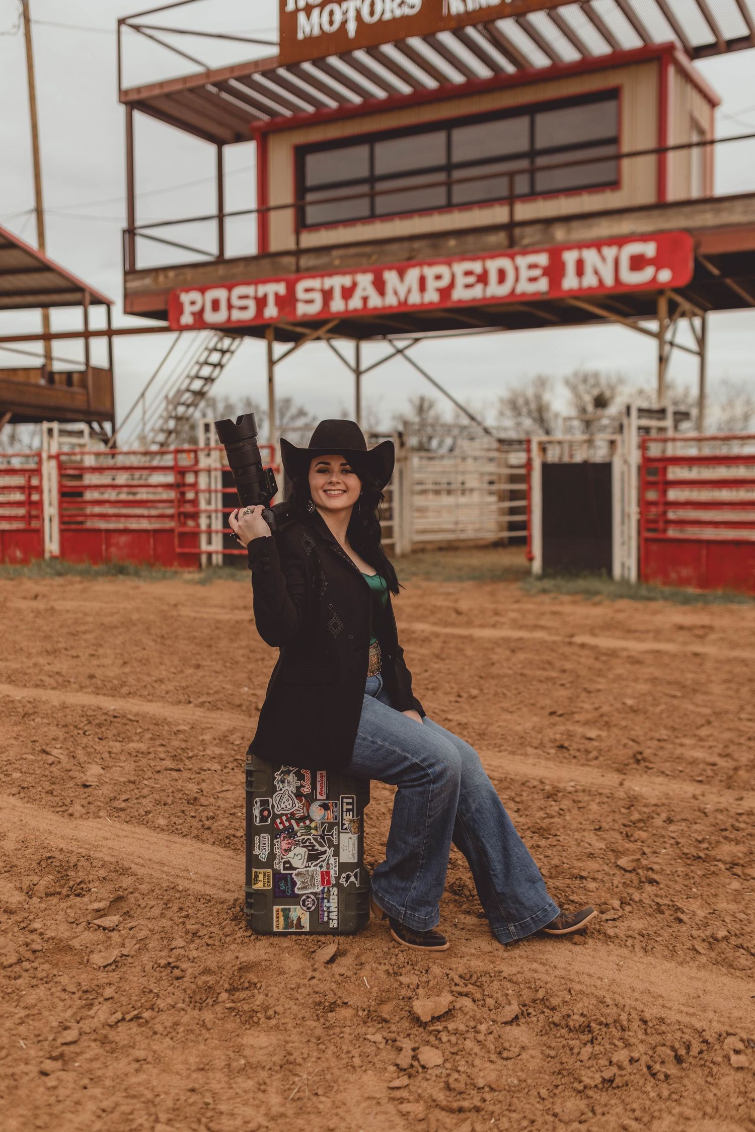 Western style photo at Post Stampede rodeo arena with figure in black jacket and jeans sitting on decorated trunk in dirt.