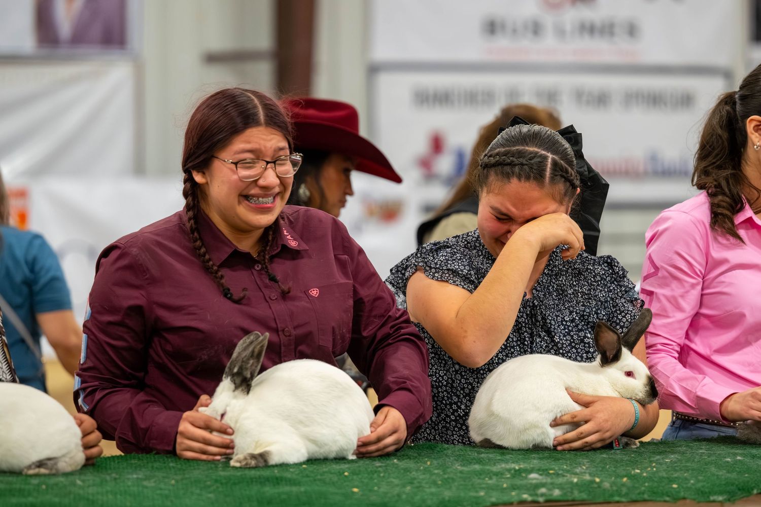 Two women smile and laugh while holding white rabbits at an indoor animal show competition.