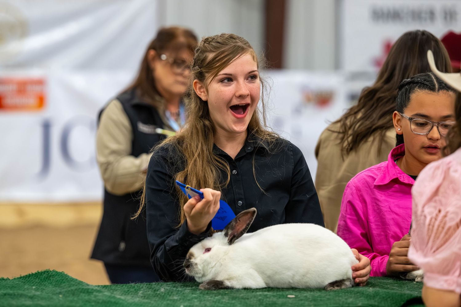 Excited girl holding a blue ribbon next to a white rabbit at a youth livestock show competition.