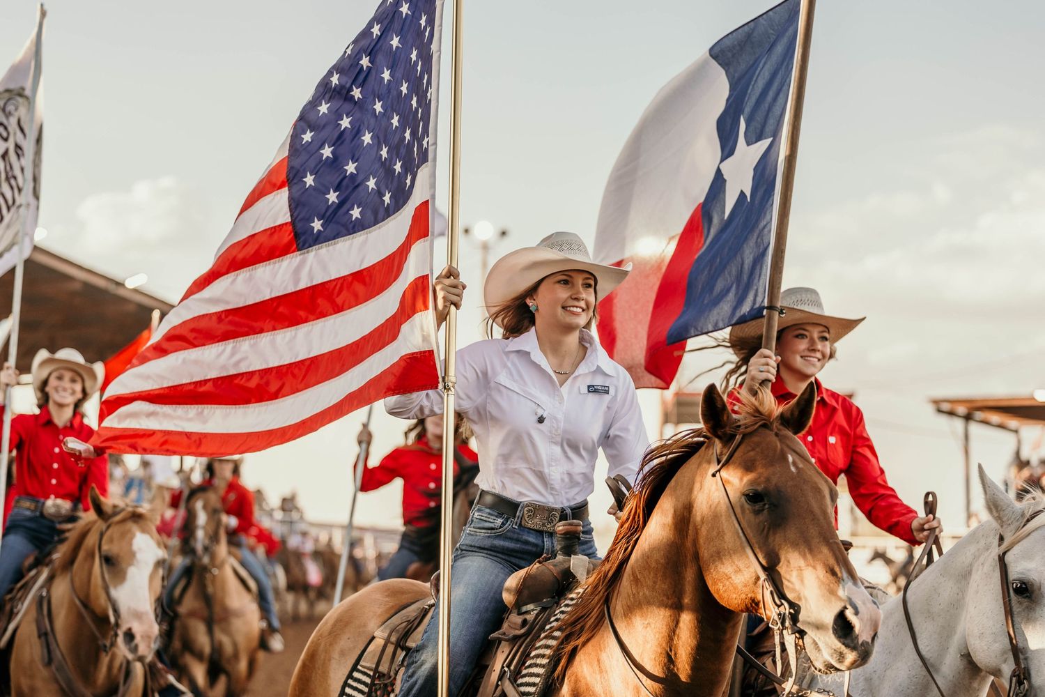 Riders carrying American and Texas flags during rodeo with crowd in background.
