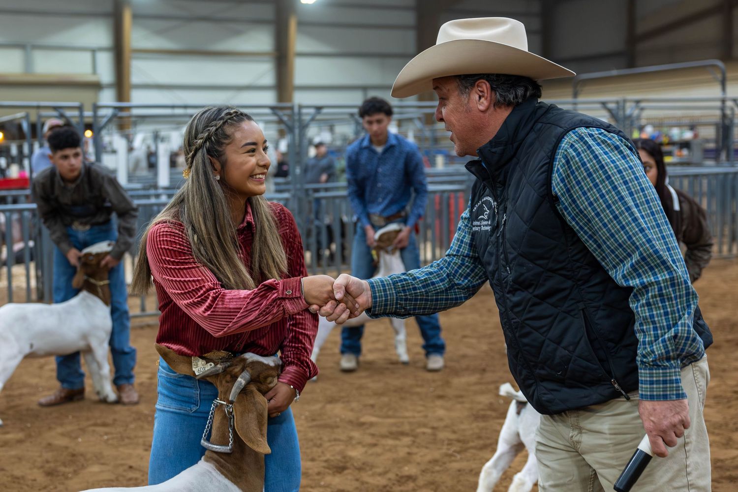 Two people shake hands at an indoor horse arena as others look on in the background.