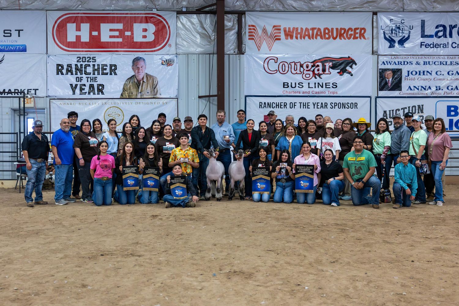 Large group of participants gathered in front of sponsors' banners at a livestock arena event.
