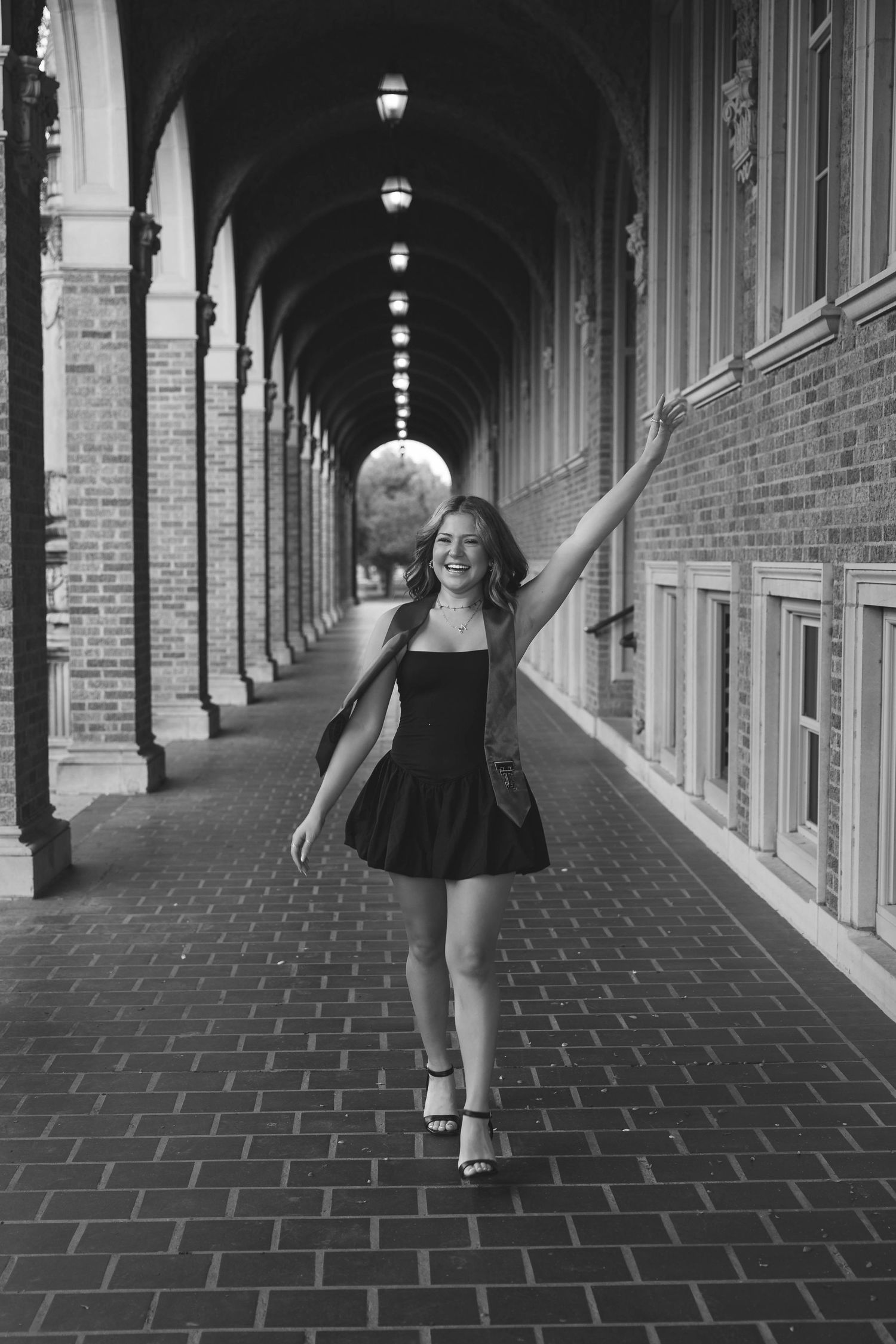 A view down a columned brick archway corridor with someone in a black dress posing joyfully.
