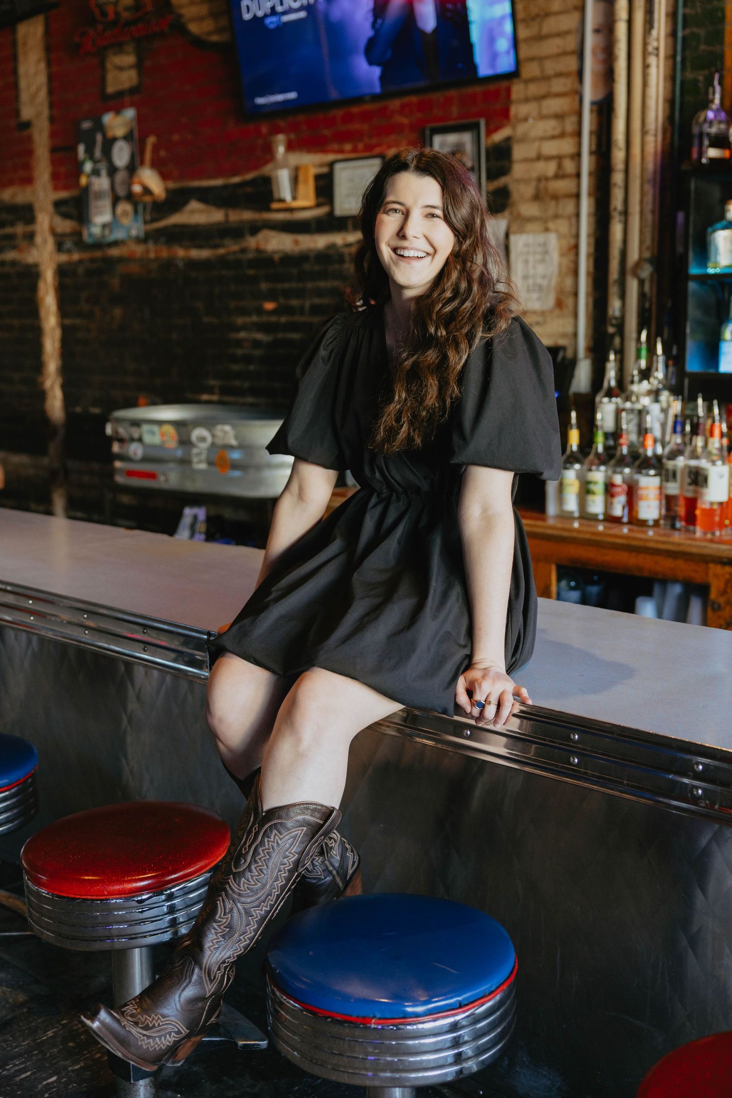 Person sitting at a bar counter wearing a black shirt and patterned pants against exposed brick walls and liquor bottles.