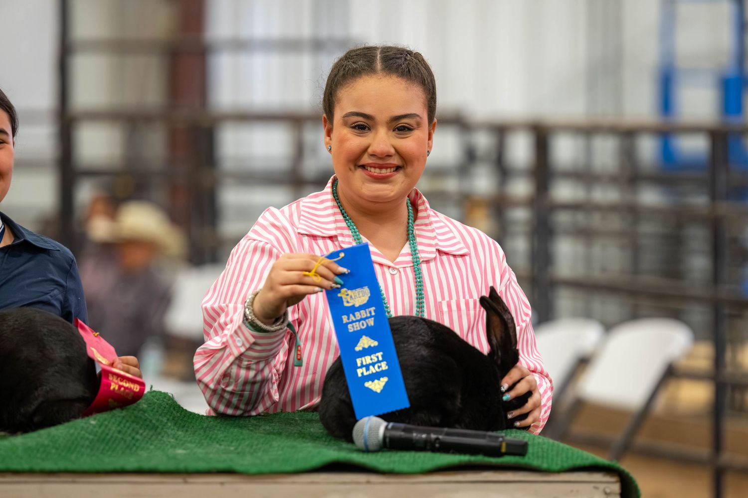 Young woman in pink striped shirt proudly holds a blue first place ribbon at an indoor livestock or county fair event.