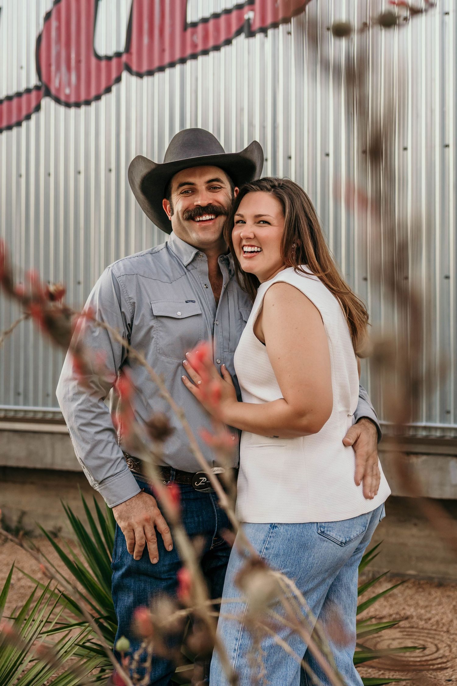 A couple in western attire embracing and smiling against a metal wall background.