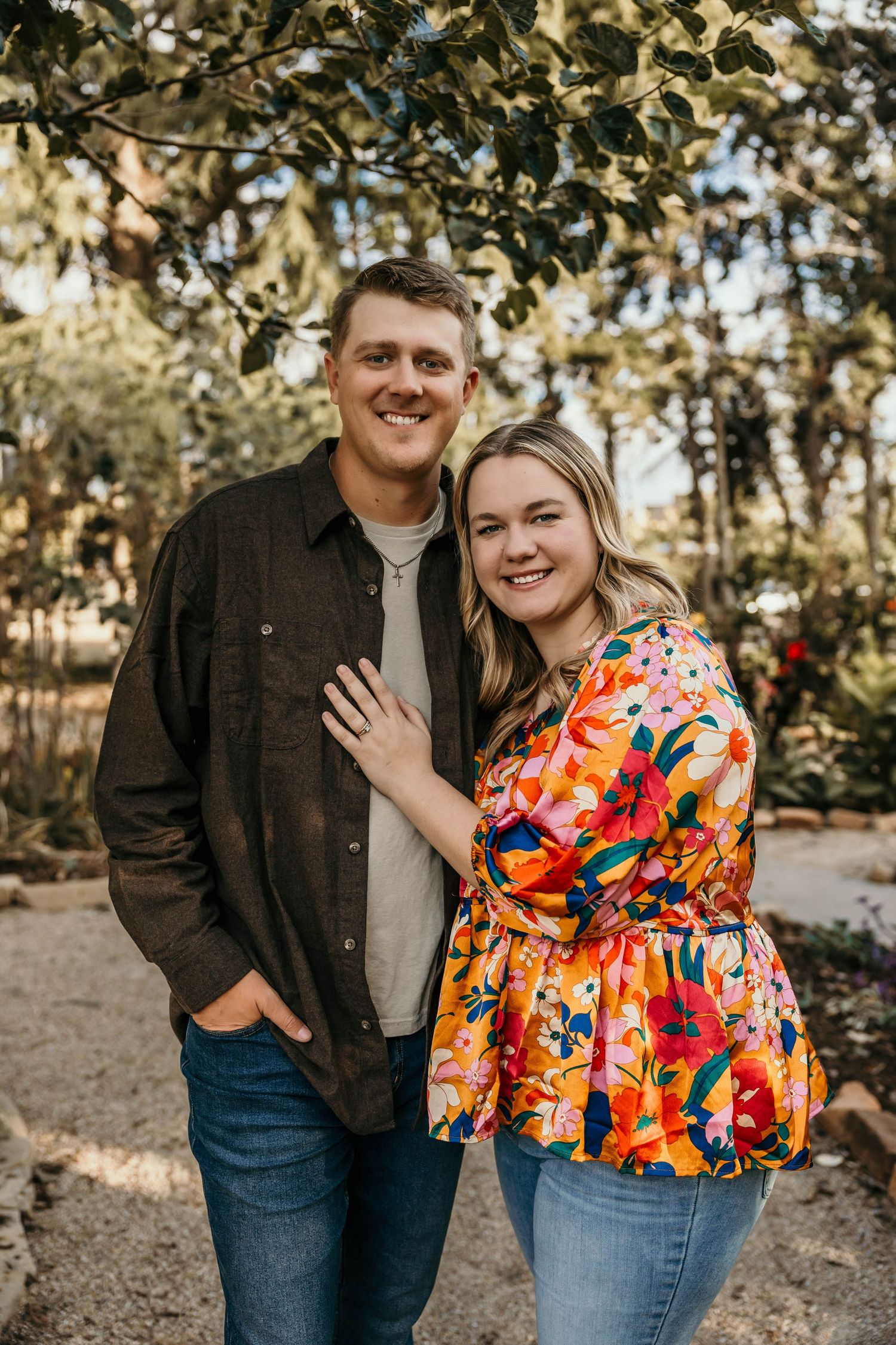 A couple in casual fall attire poses for an engagement photo under tree branches.