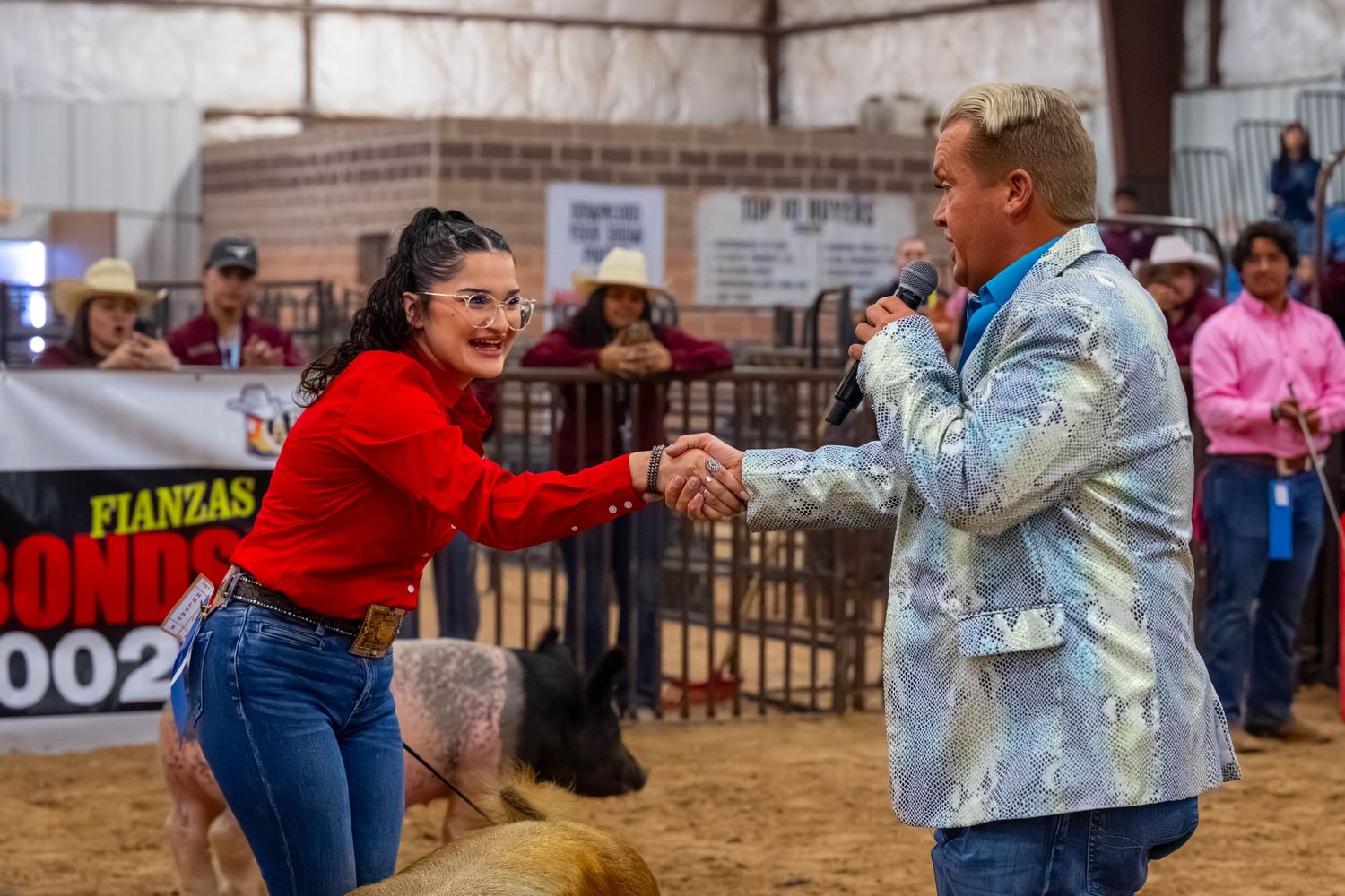 A woman in a red top shakes hands with a man holding a microphone at a livestock auction.