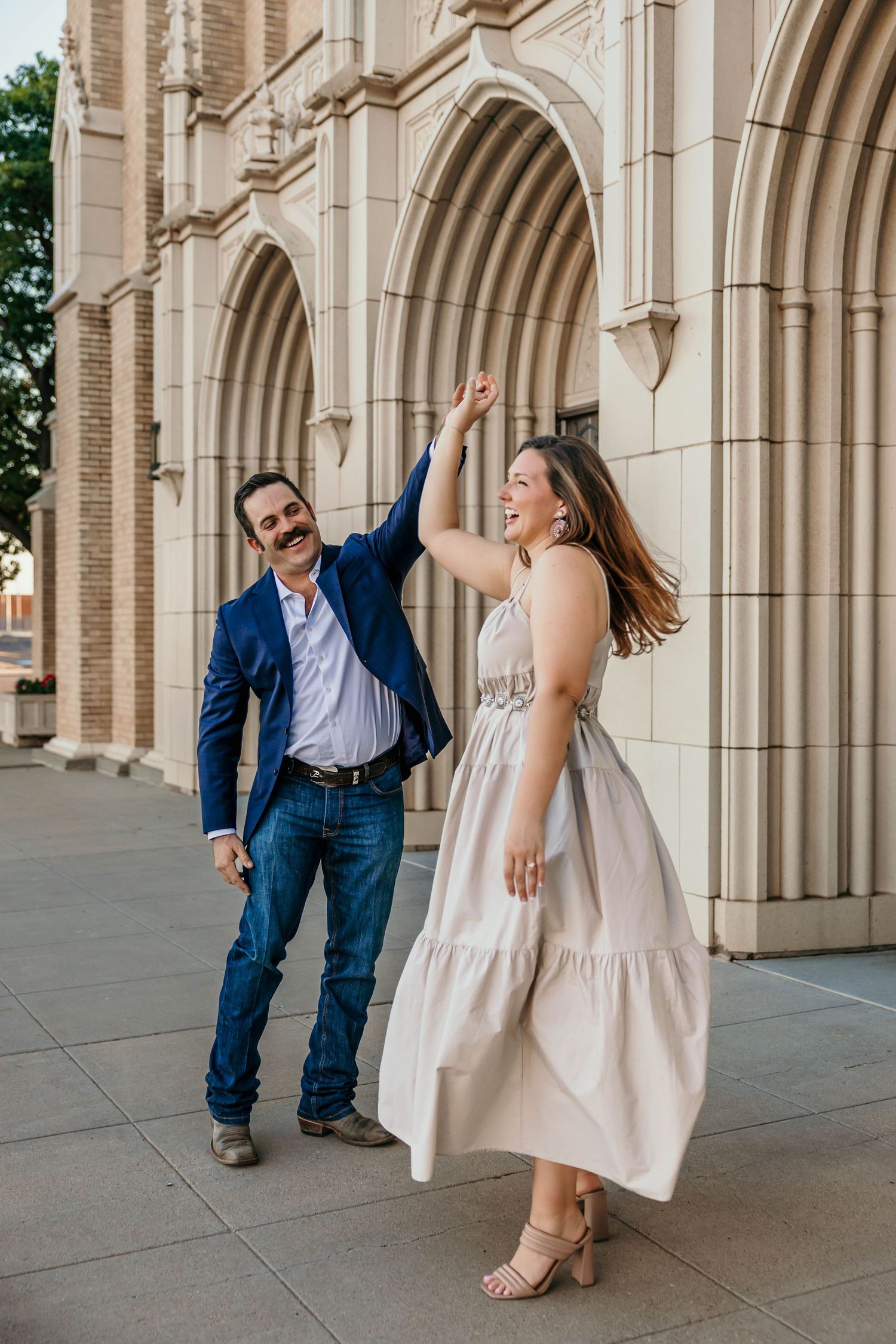 A couple shares a playful dance moment under an ornate stone archway of a historic building.
