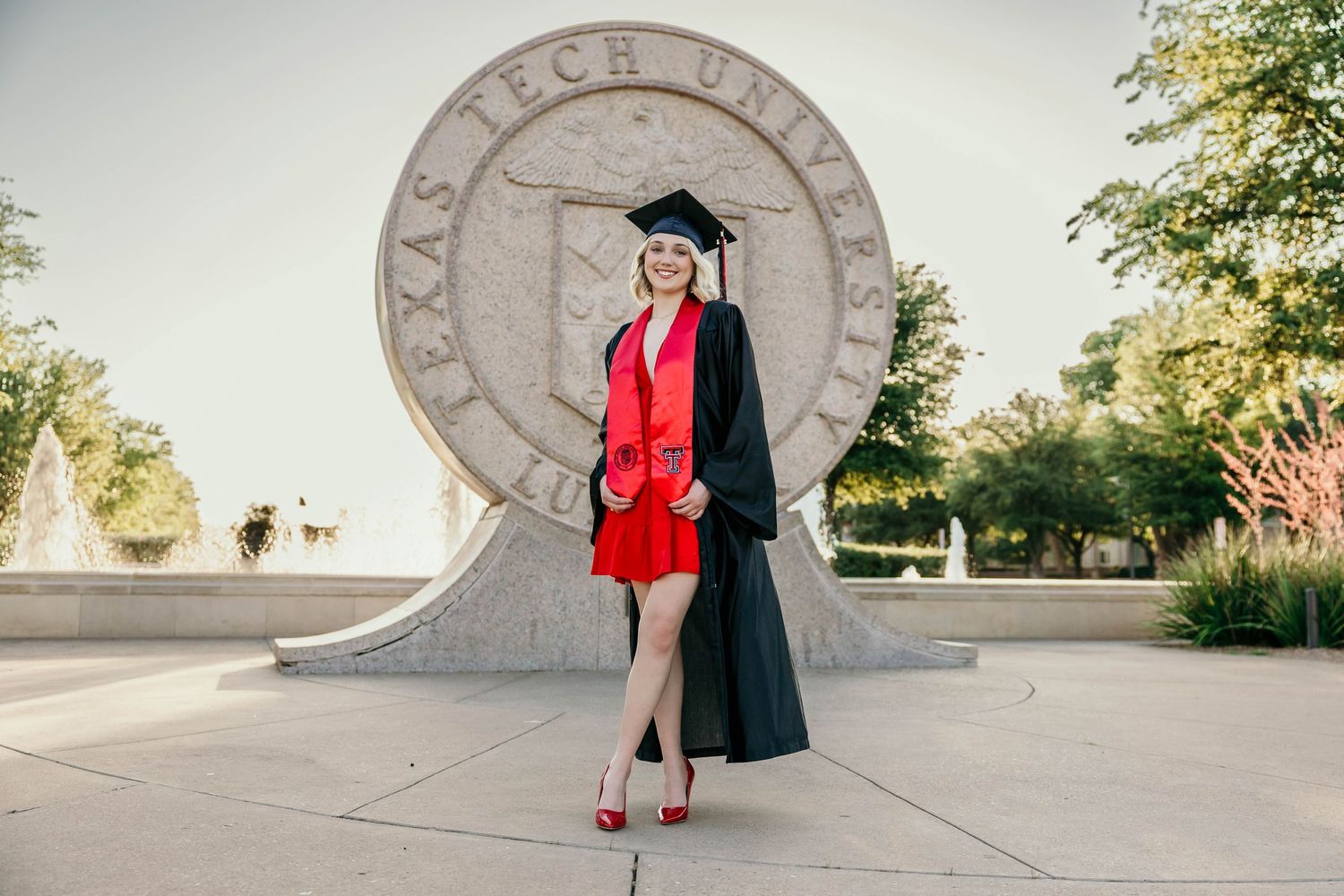 Graduate in red dress and black cap and gown stands in front of Texas Tech University seal on sunny day.