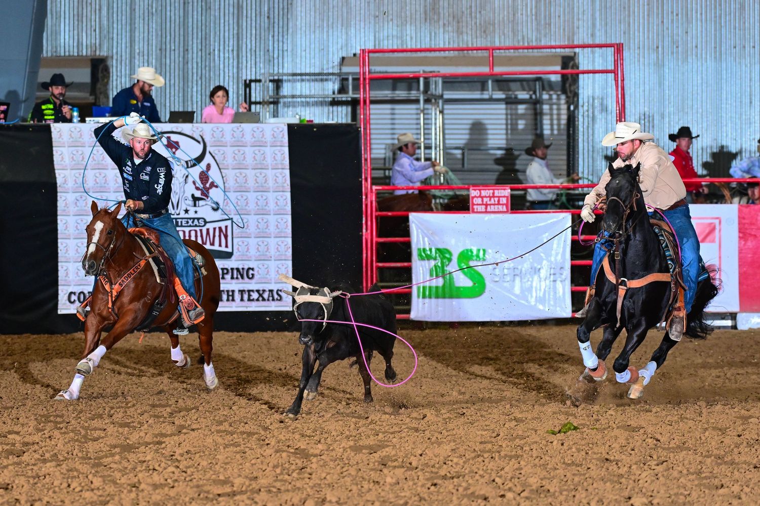 Two cowboys on horseback compete in a team roping event at an indoor Texas rodeo arena.