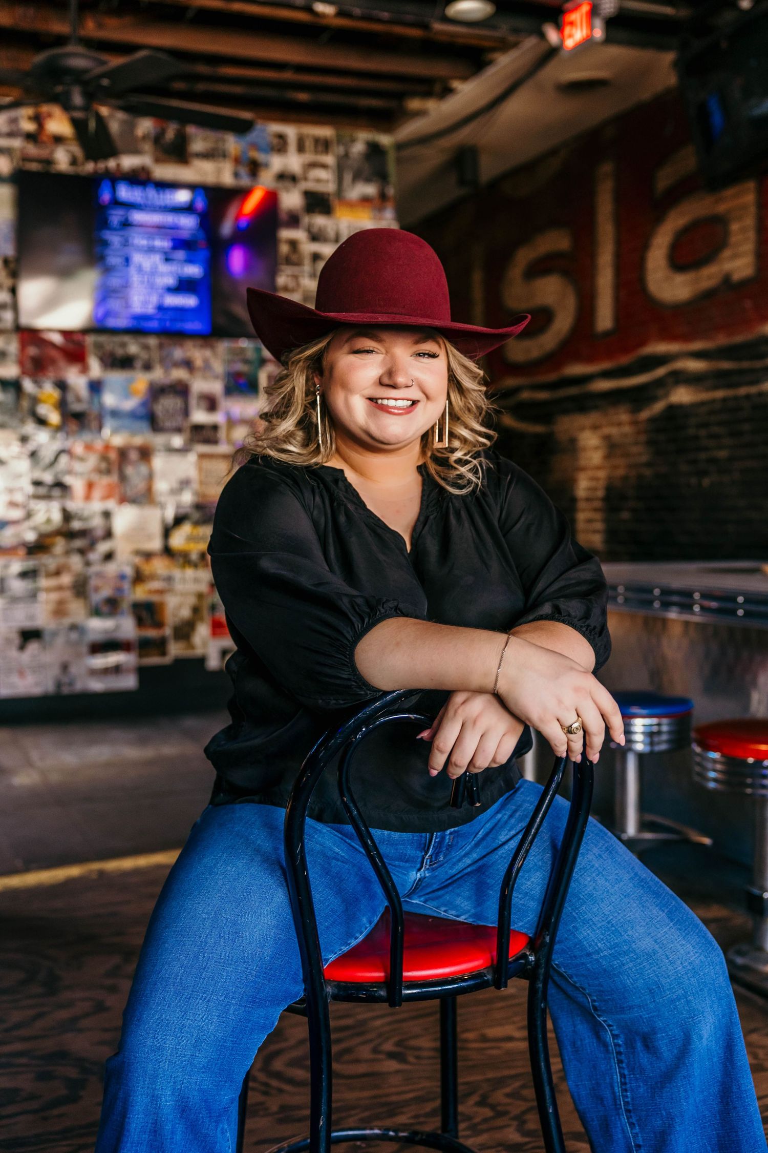 A person wearing blue jeans and a black top sits casually on red chairs in a dark, brick-walled venue.