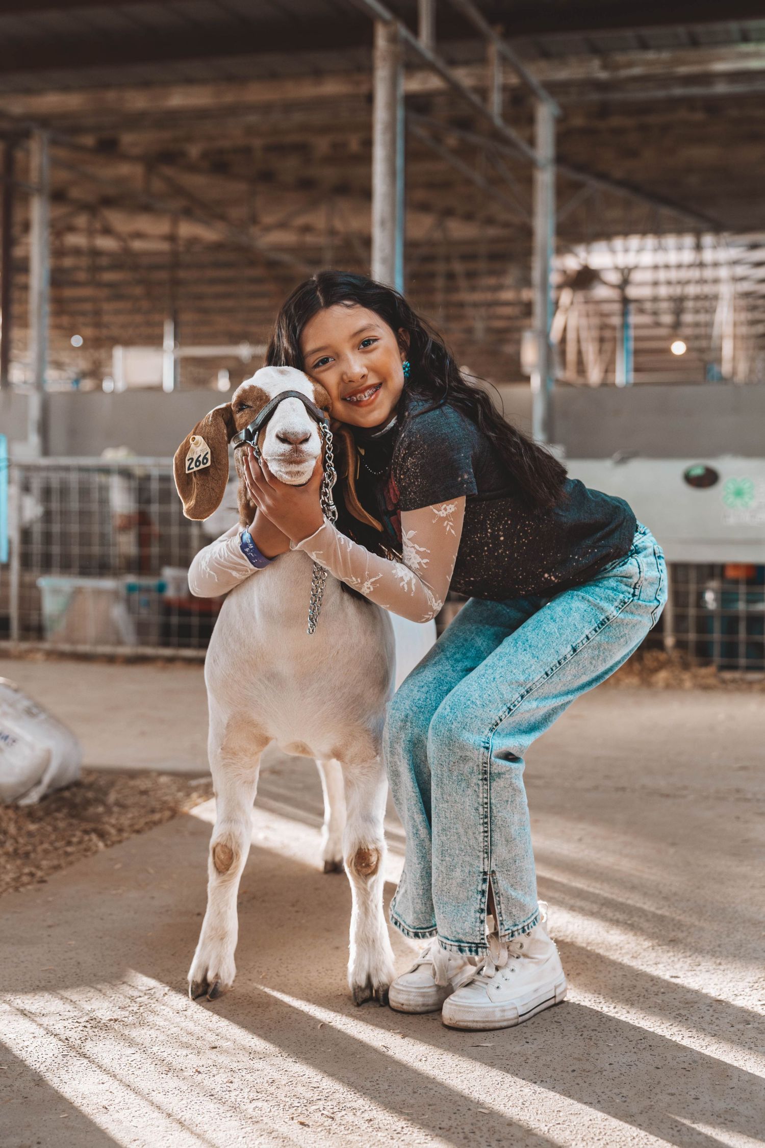 A person in a black top and light jeans crouches next to a smiling white goat in a rustic barn setting.