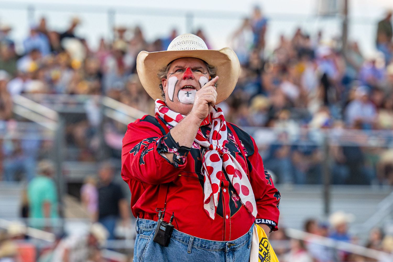 Rodeo clown in red shirt, cowboy hat, and face paint performs for a crowd at a western rodeo event.