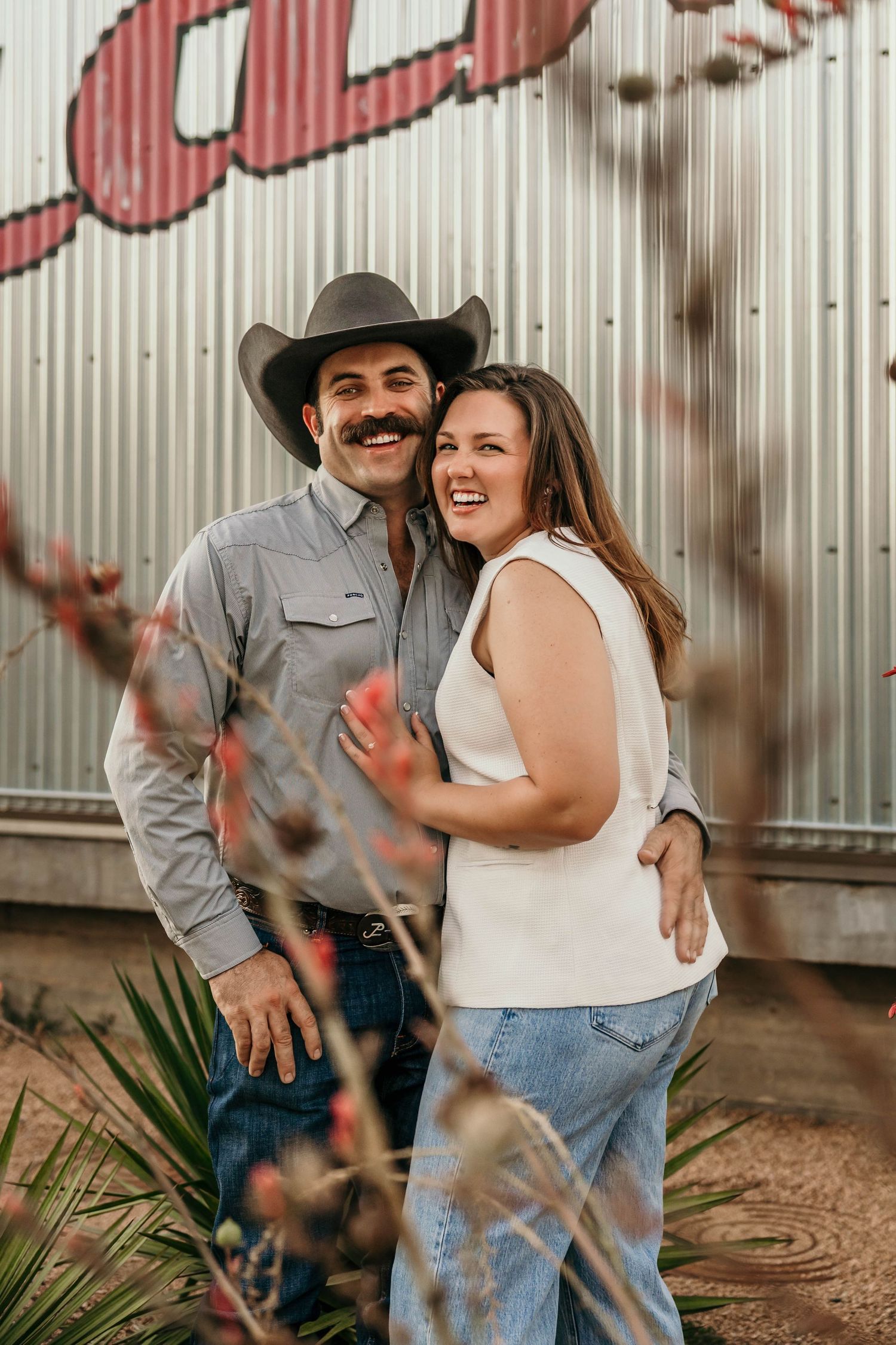 A couple in casual western attire share romantic moments against a rustic corrugated metal wall backdrop.