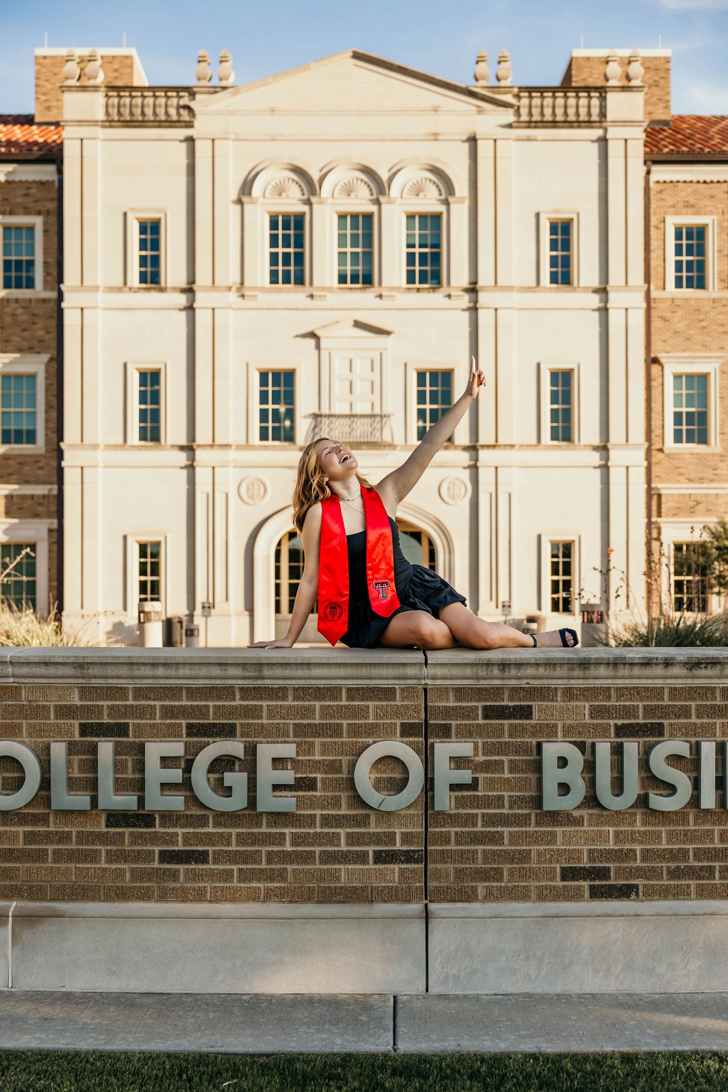 Graduate poses in red blazer atop brick wall outside College of Business building at sunset.