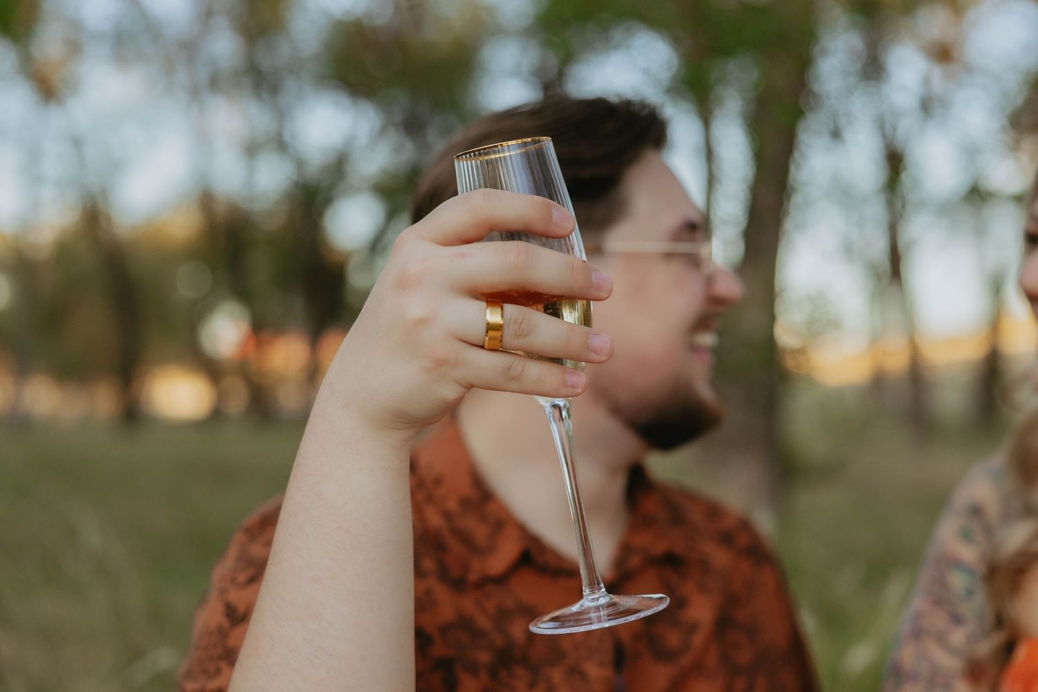 A person holding a wine glass outdoors at sunset in a natural setting with warm lighting.
