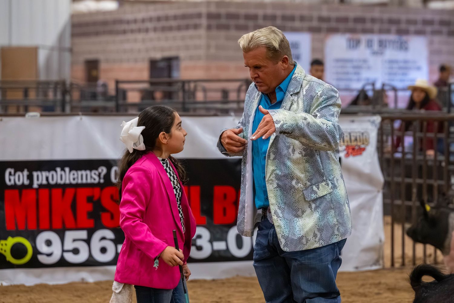 Man in blue jacket talks with young girl in pink at an indoor rodeo arena with sponsor banners.