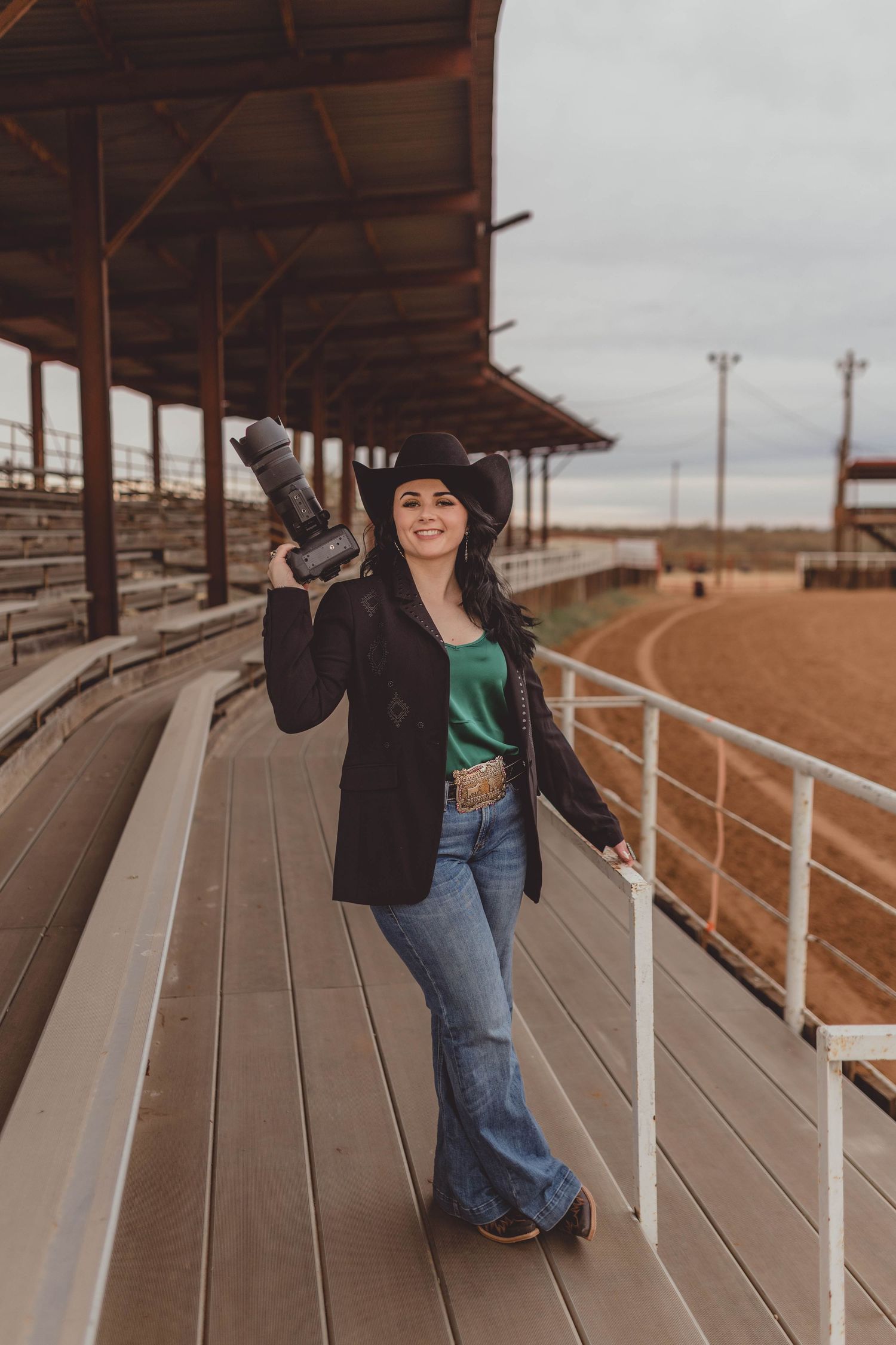 Woman in cowboy hat holding camera at rodeo arena bleachers, wearing jeans and green top.