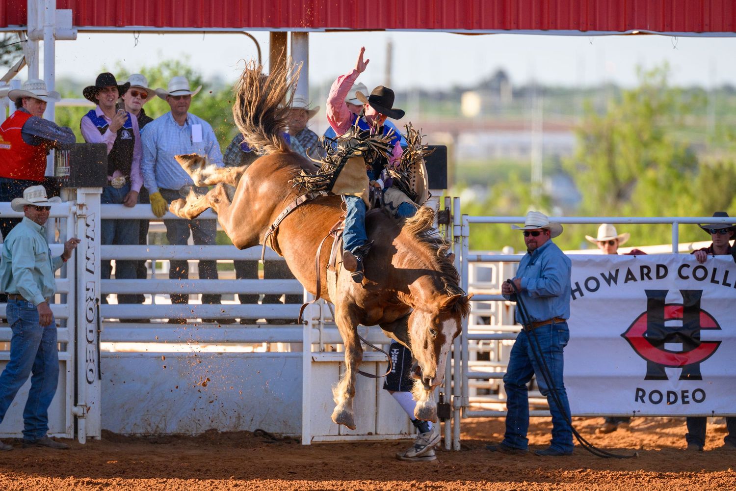 Cowboy rides bucking bronco at Howard College Rodeo event with spectators watching from arena fence.