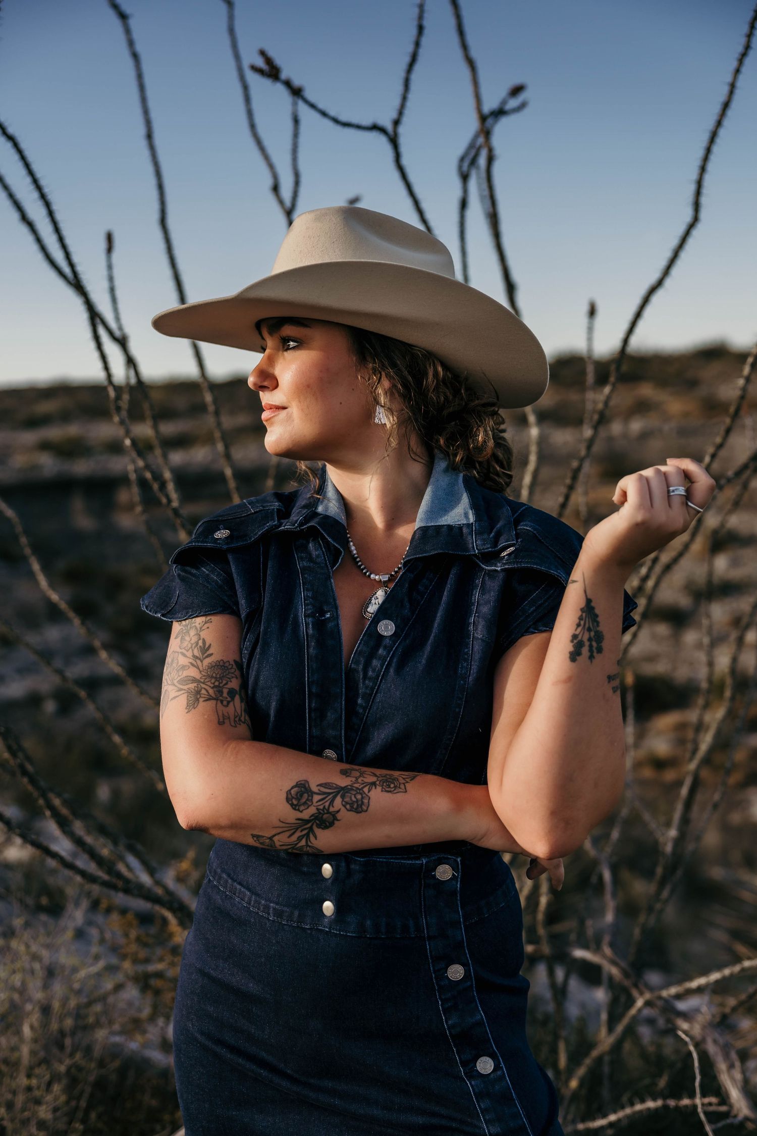 A denim-clad figure with visible tattoos and a tan cowboy hat poses outdoors against desert brush at sunset.