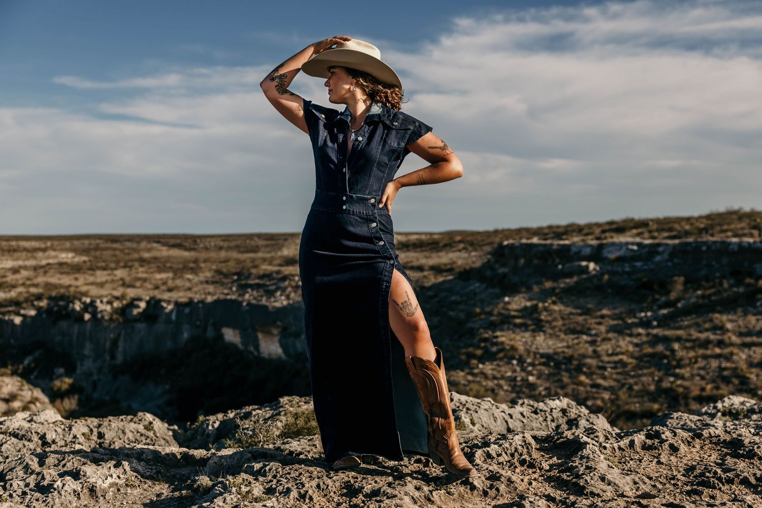 A stylish figure in a black dress and cowboy hat poses dramatically on rocky terrain against a blue sky.