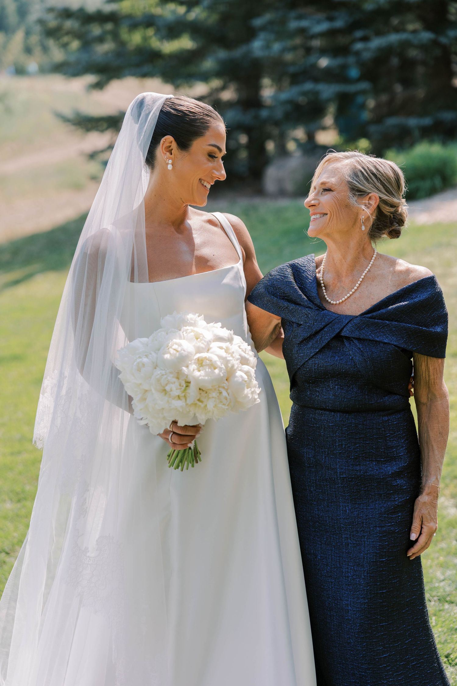 A bride in white gown and bridesmaid in navy dress share joyful candid moments together outdoors.