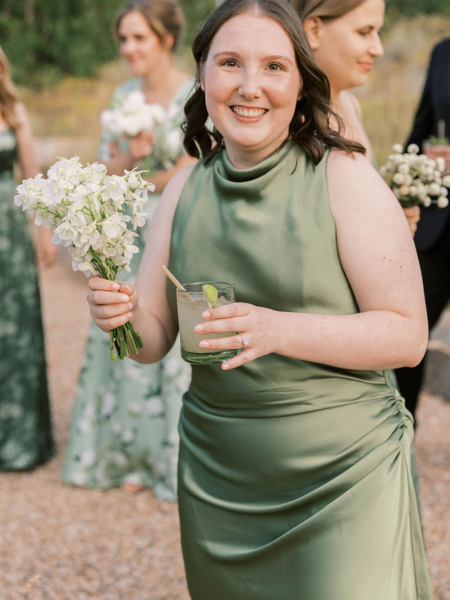 Bridesmaids in sage green dresses hold delicate white bouquets during an outdoor wedding ceremony.