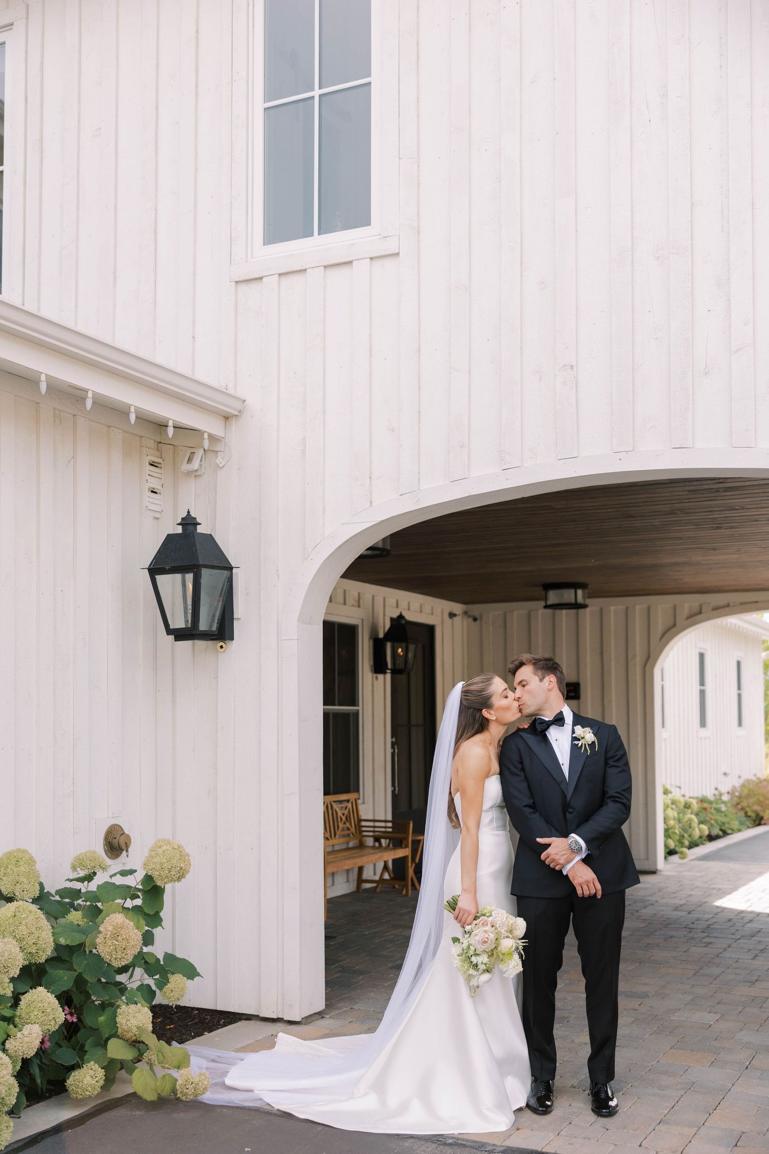 Wedding couple posing outside white barn-style venue with romantic architectural details.