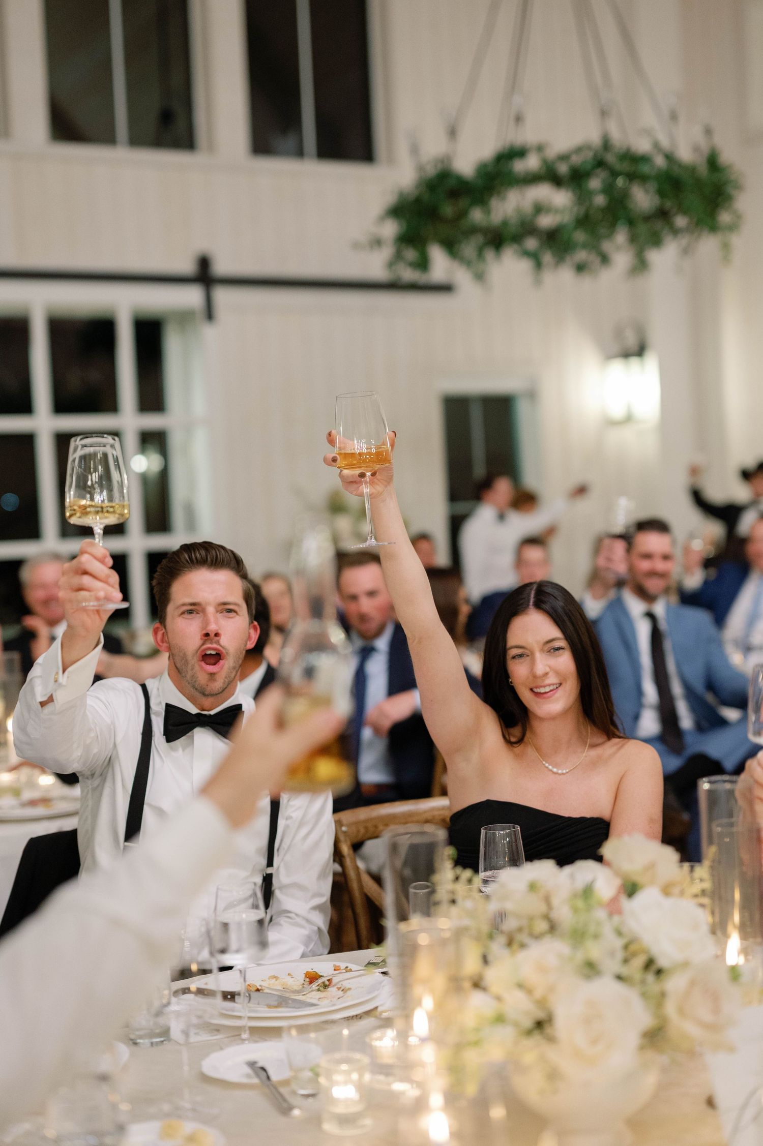 Wedding guests celebrate joyfully at an elegant reception dinner with white floral centerpieces and candlelight.