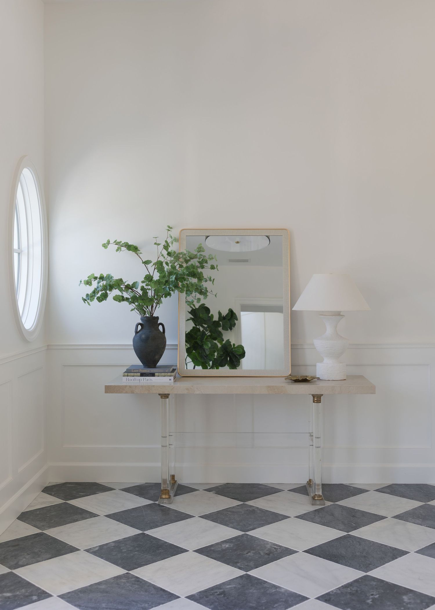 Elegant entryway with checkered marble floors, acrylic console table, square mirror, white lamp, and dark vase with greenery.
