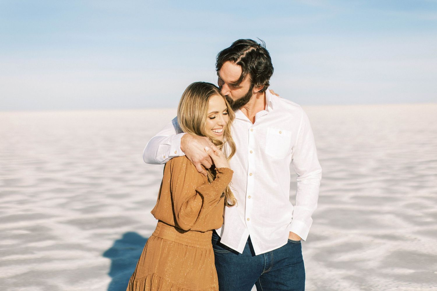 Couple in white and tan clothing embracing on crystalline salt flats with blue sky backdrop.