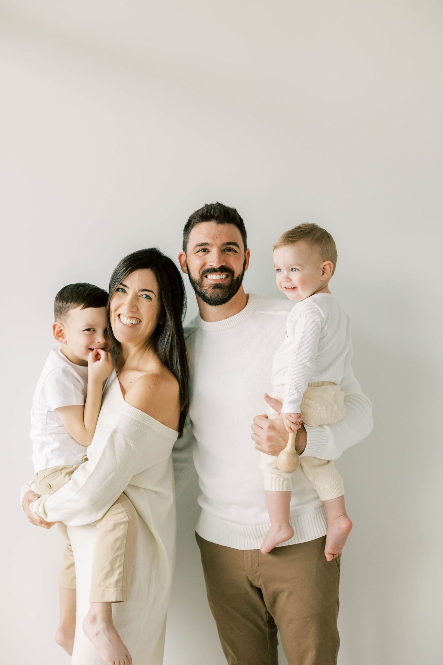 Happy family of four wearing coordinated white outfits pose against light wall.