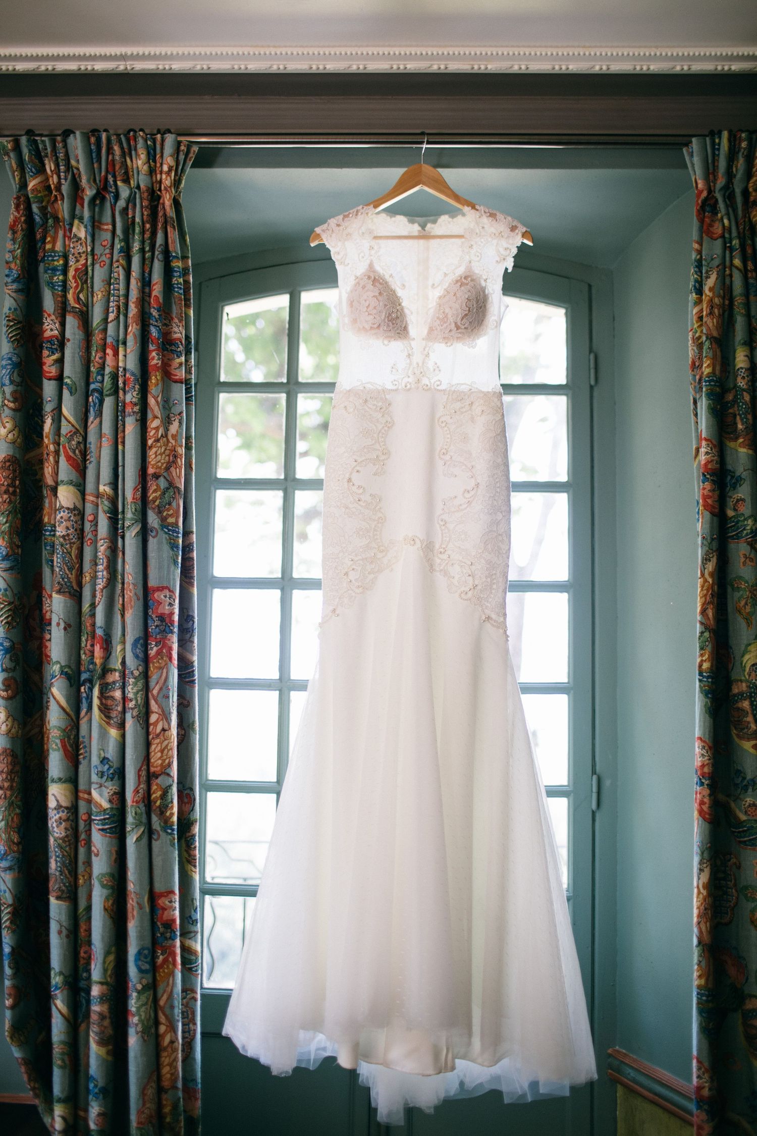 Elegant white lace wedding dress hanging in window with floral curtains.