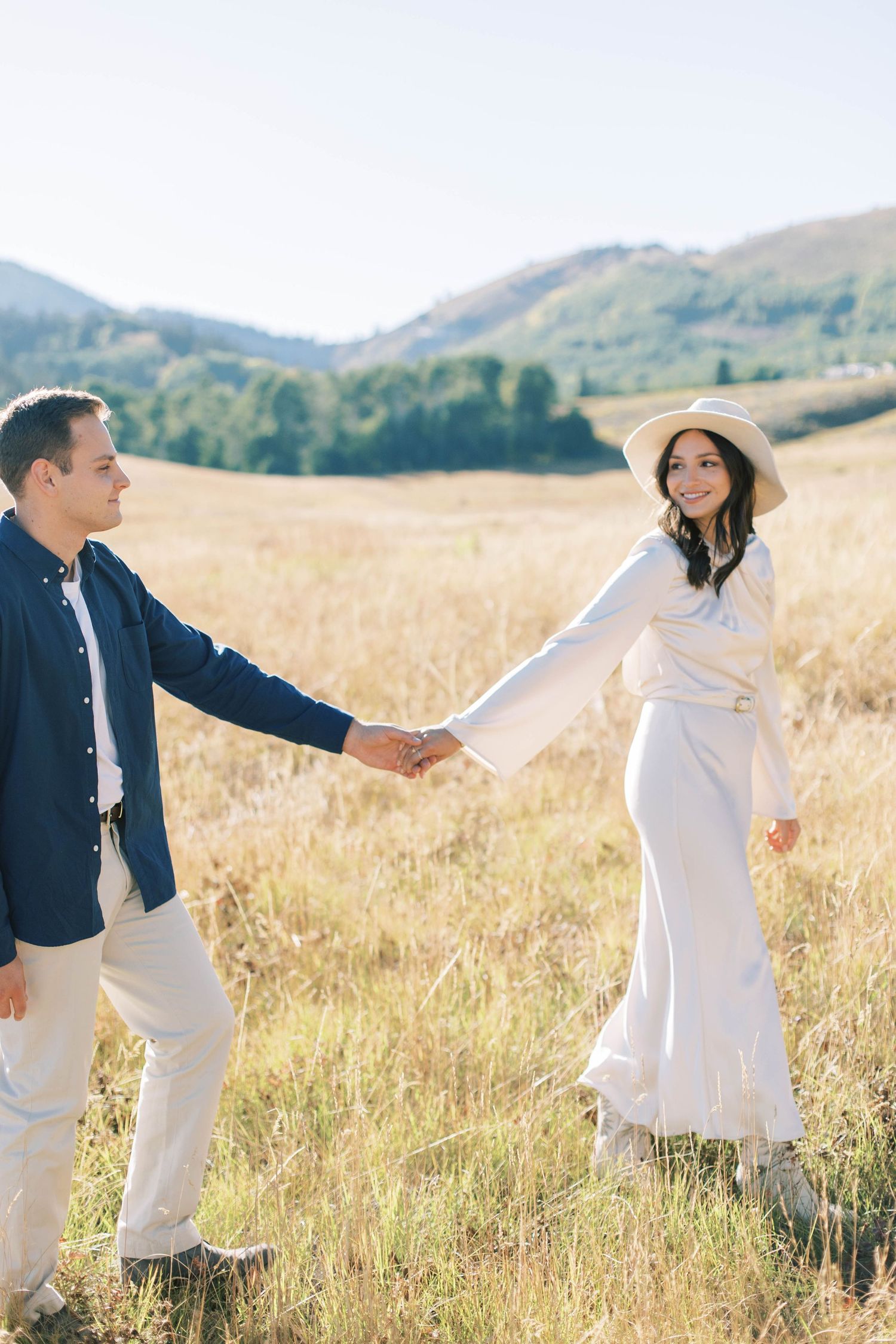 Couple holding hands in a golden field with mountains in the background wearing coordinating white and navy outfits.