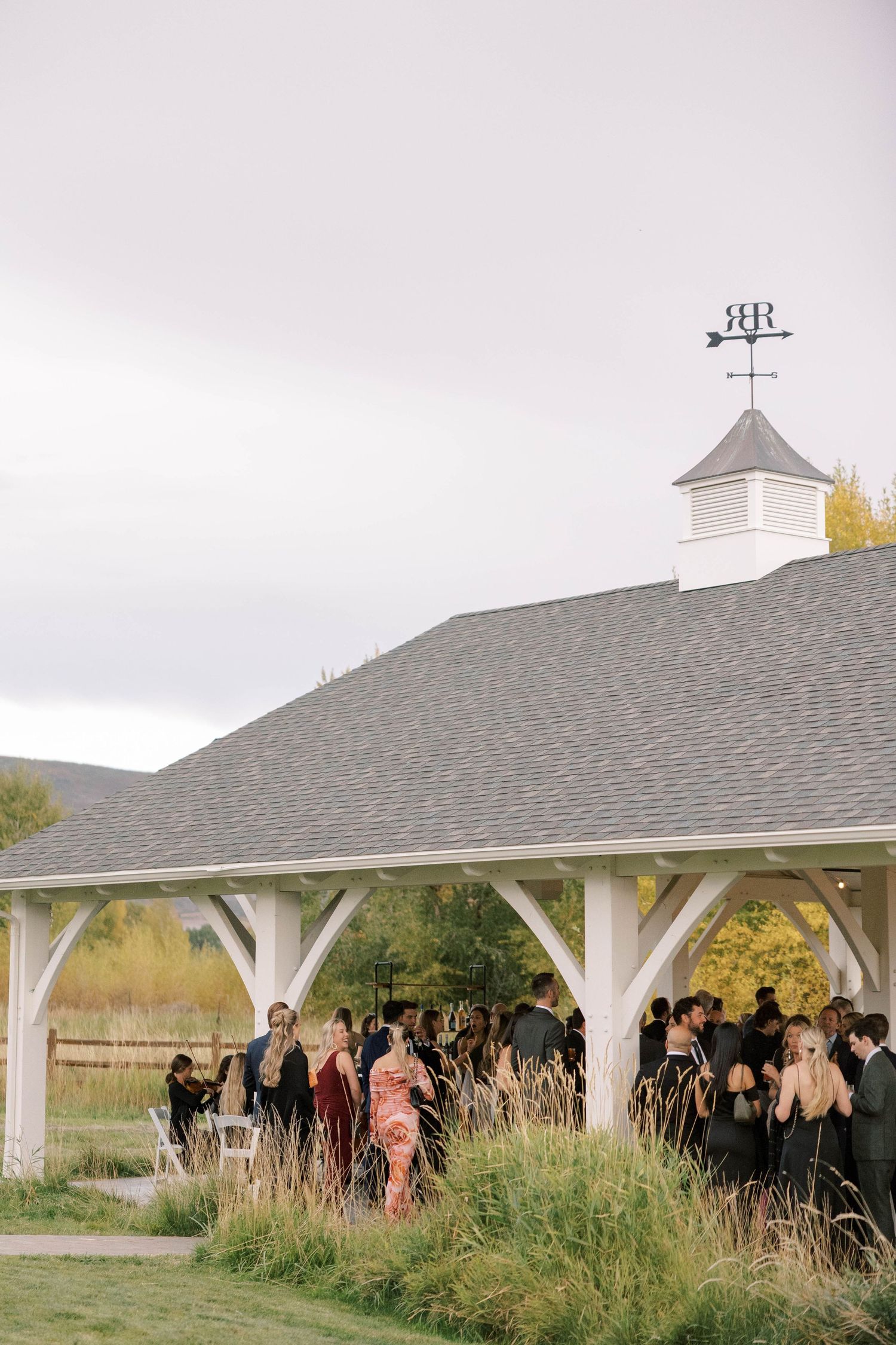 Wedding guests gather under a white pavilion with autumn foliage visible in the background.