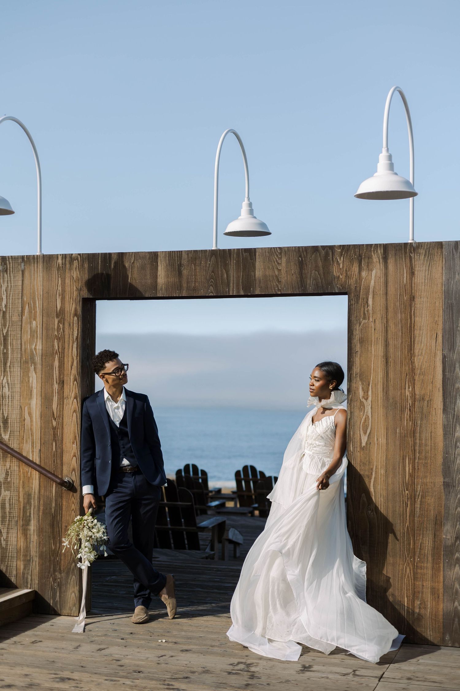A romantic wedding photoshoot on a rustic wooden deck overlooking the ocean with pendant lights and coastal scenery.