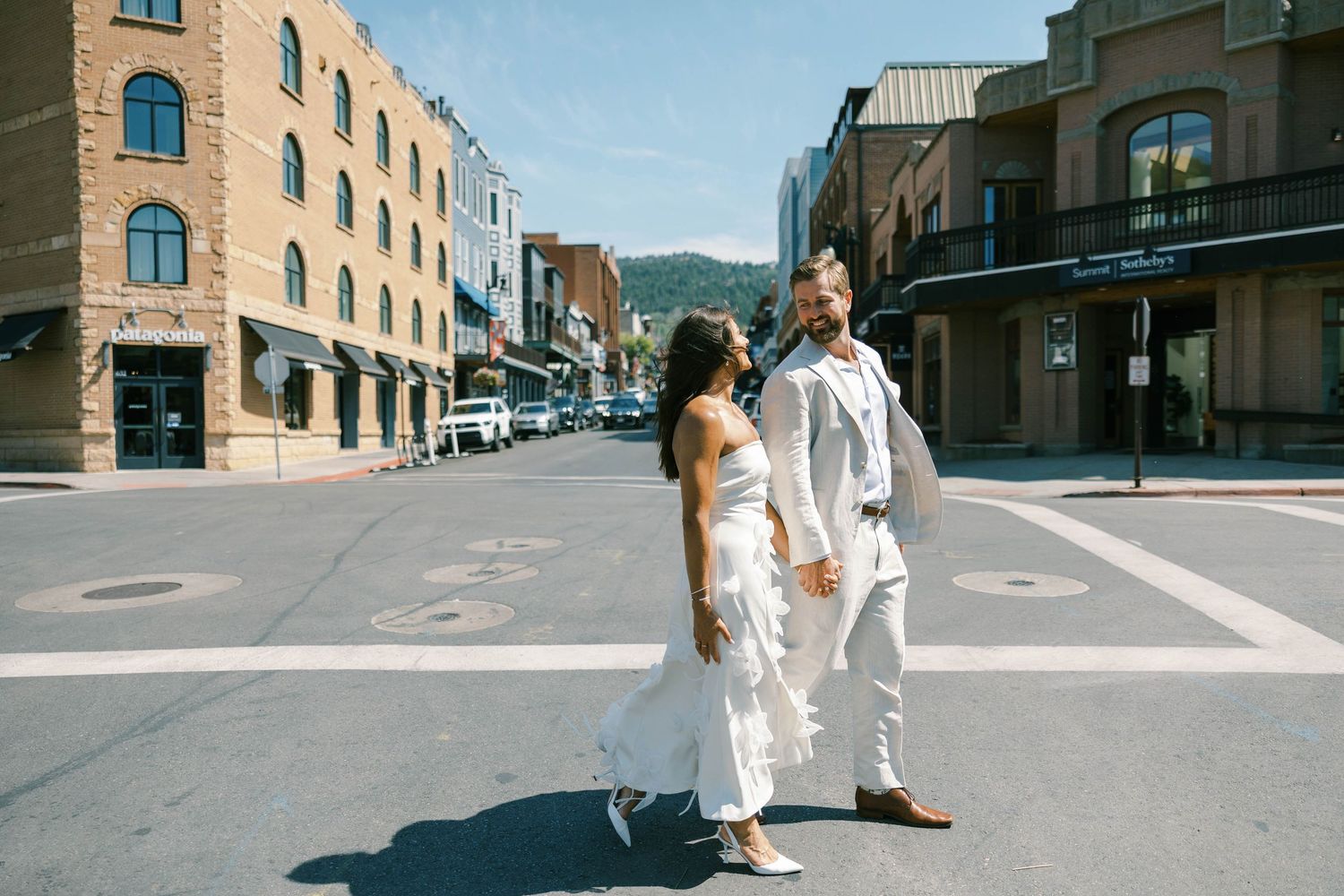 A couple in white outfits poses in the middle of a city street with brick buildings and mountains in the background.