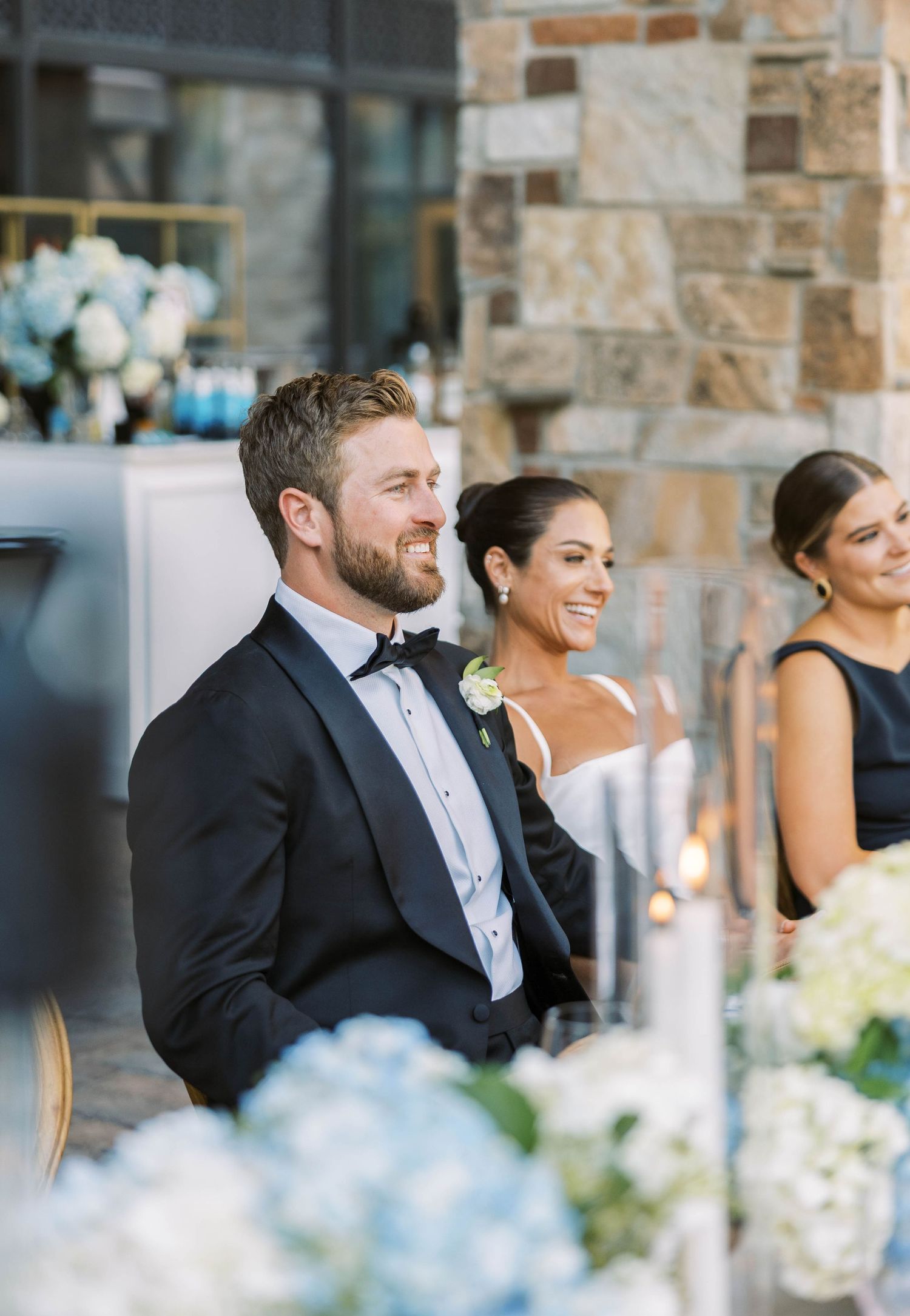 Wedding party members laugh together while seated at an elegant stone venue with soft blue floral decor in foreground.