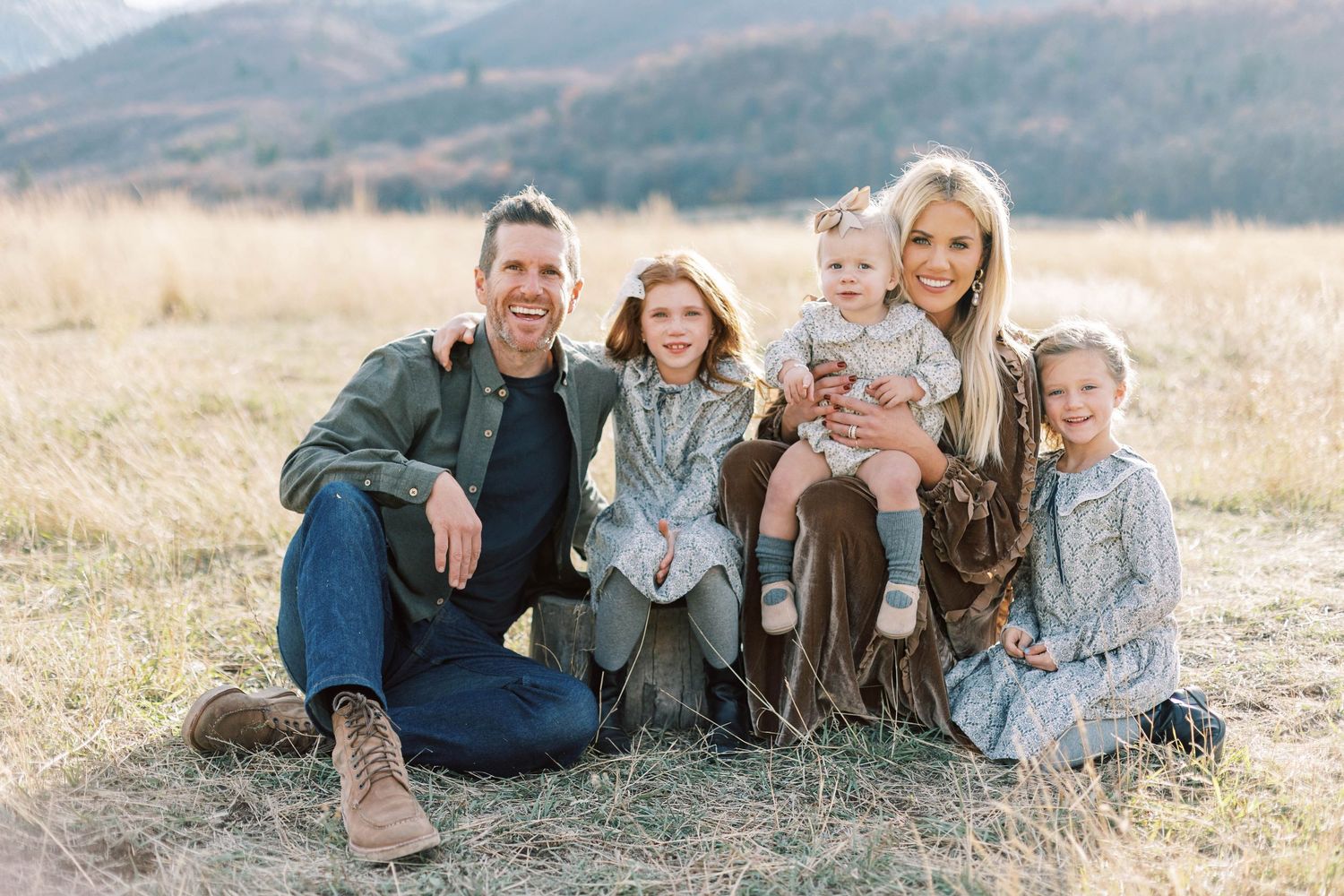 A family portrait in a sunlit Utah mountain meadow with golden grass and mountain backdrop.