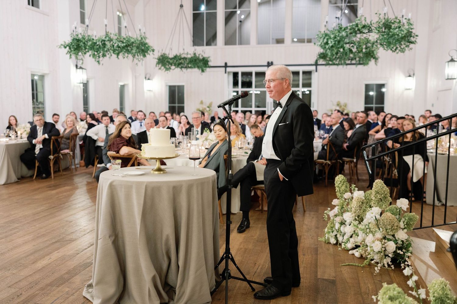 A speaker addresses guests at an elegant wedding reception in a bright white room with hardwood floors and floral arrangements.