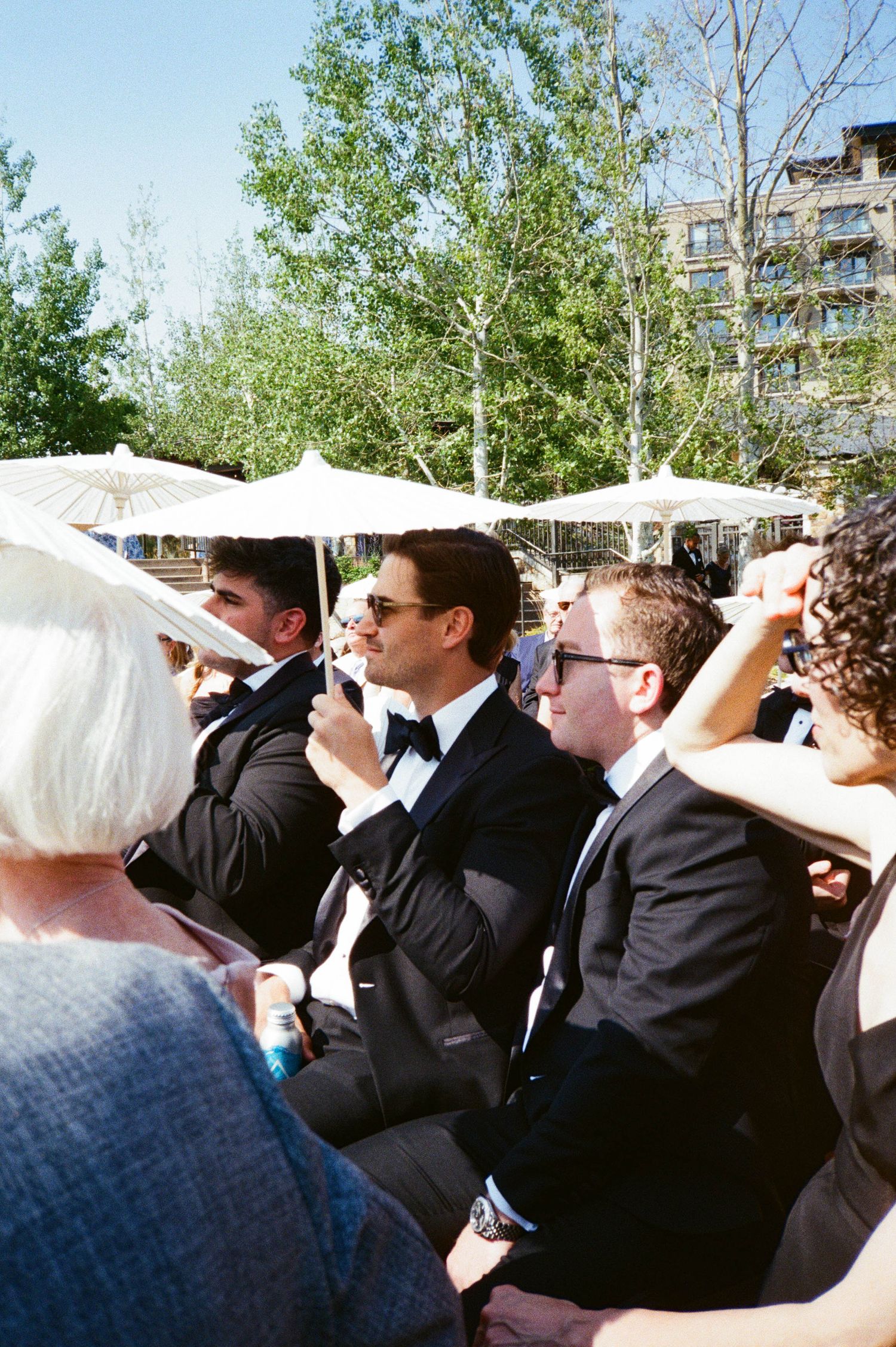 A group of formally dressed guests sit outdoors under white umbrellas at an elegant event.