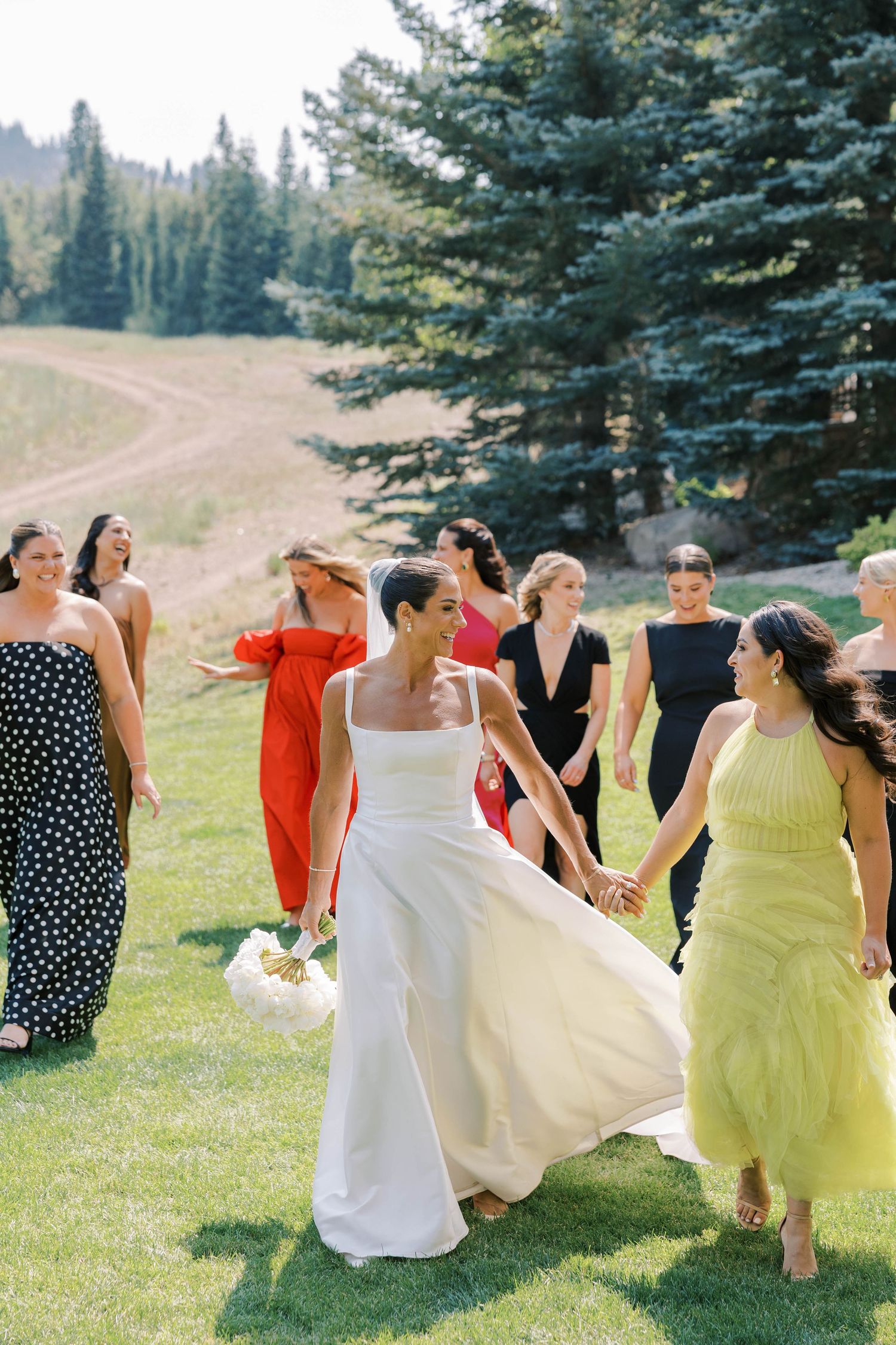 A group in yellow and white dresses dances joyfully on a grassy hillside with evergreen trees in the background.
