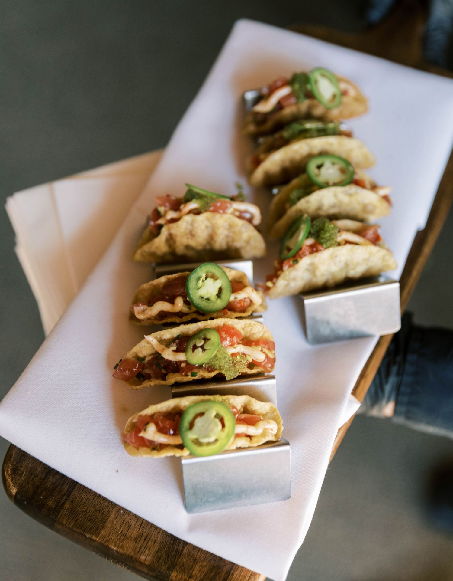 Small crispy tacos topped with fresh jalapenos and garnishes arranged on white paper on a wooden serving board.