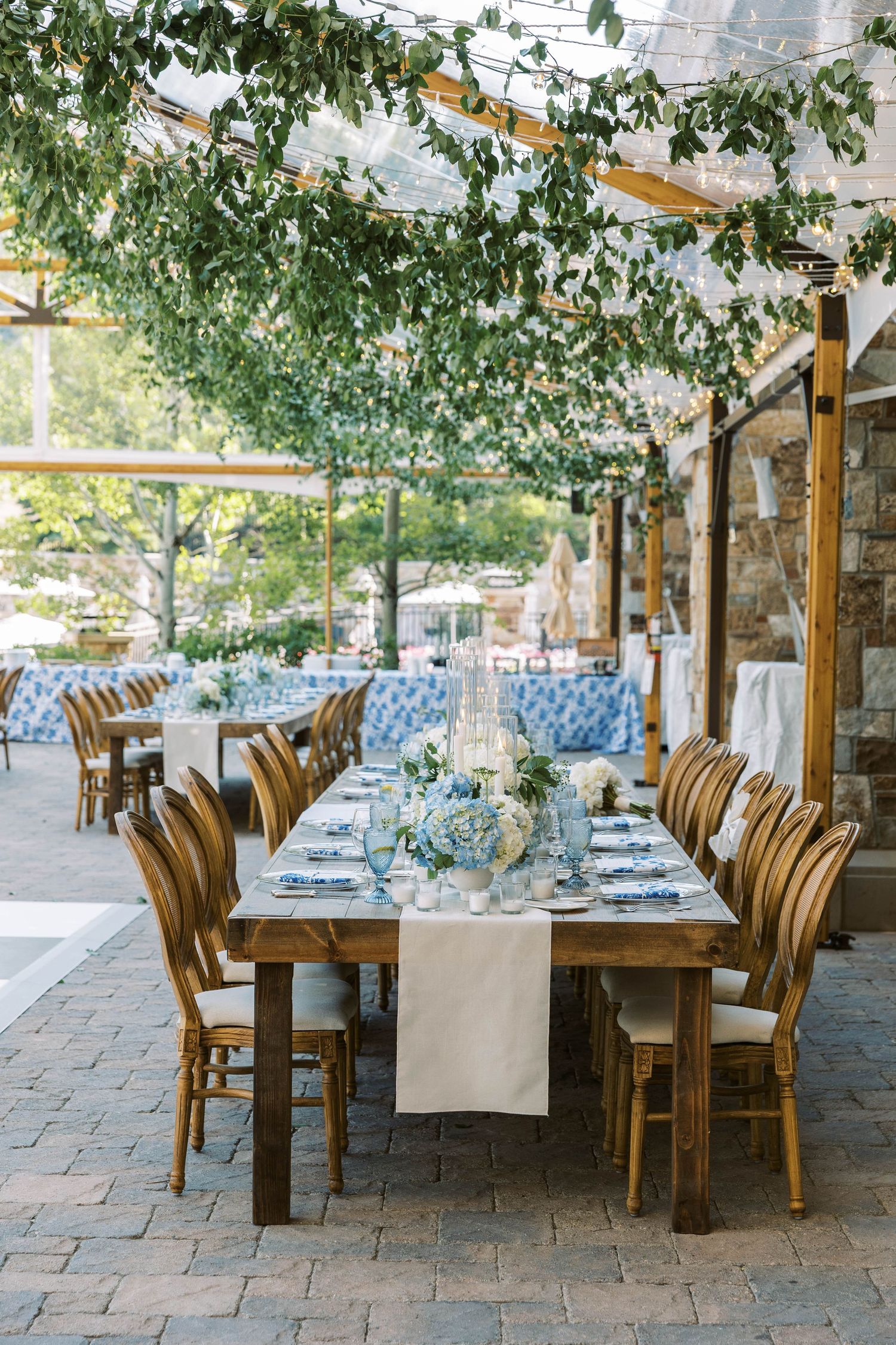 Outdoor dining setup with wooden chairs and blue cushions arranged along stone patio under leafy tree canopy.