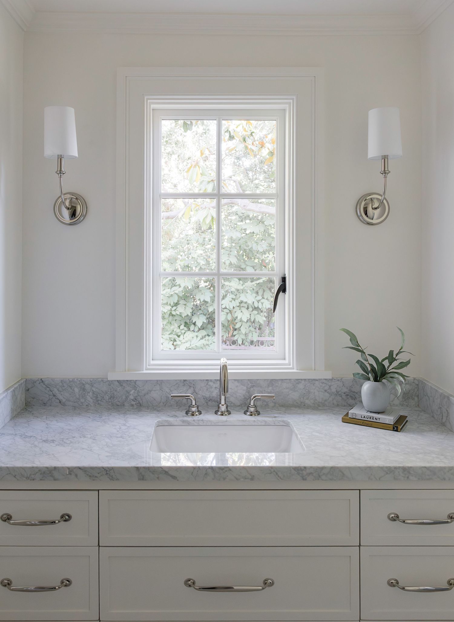 Elegant bathroom vanity with gray marble countertop, farmhouse sink, chrome fixtures, white cabinetry, and wall sconces flanking a window.