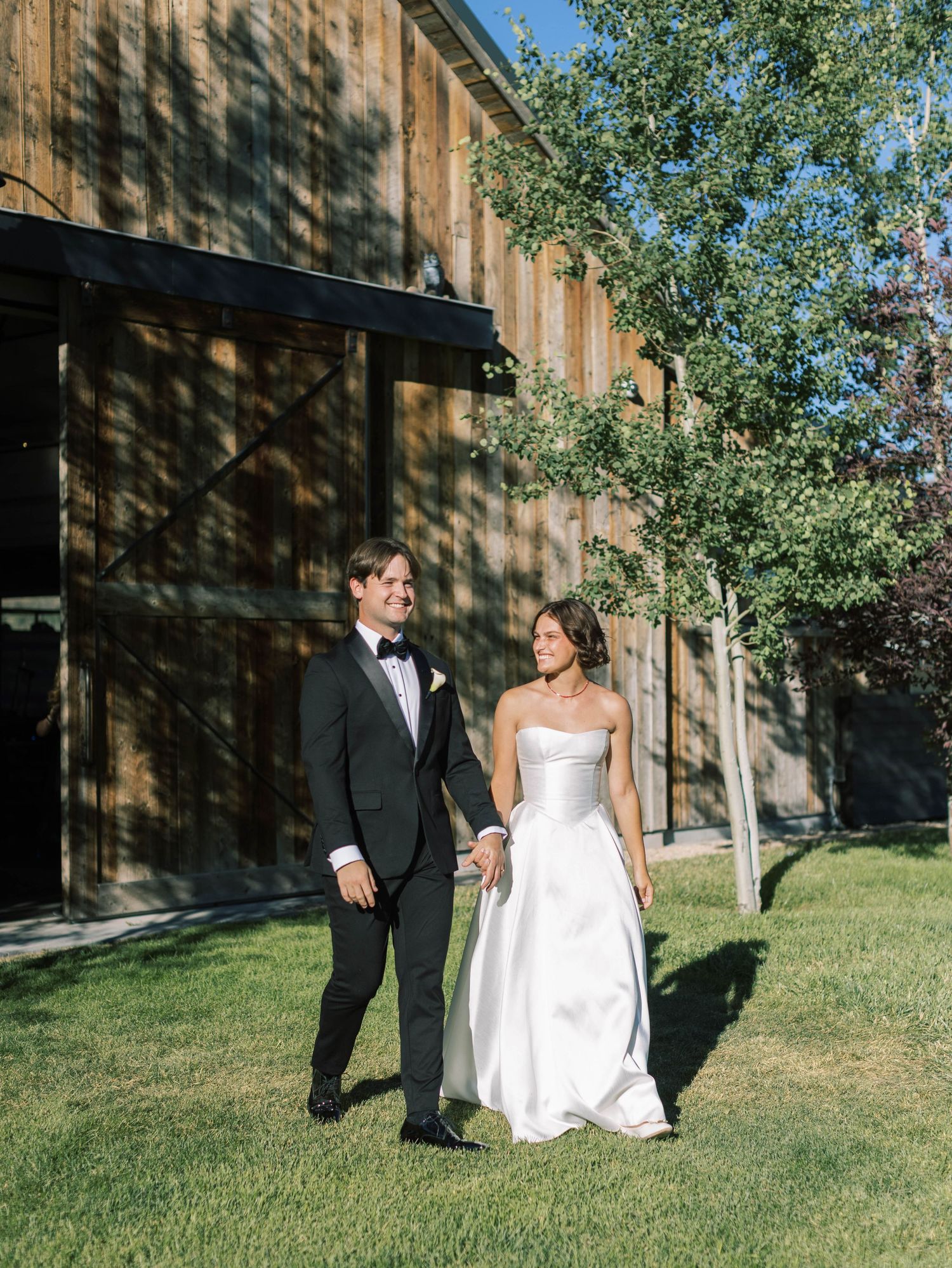 A couple in wedding attire stands together on a lawn beside a rustic wooden barn on a sunny day.