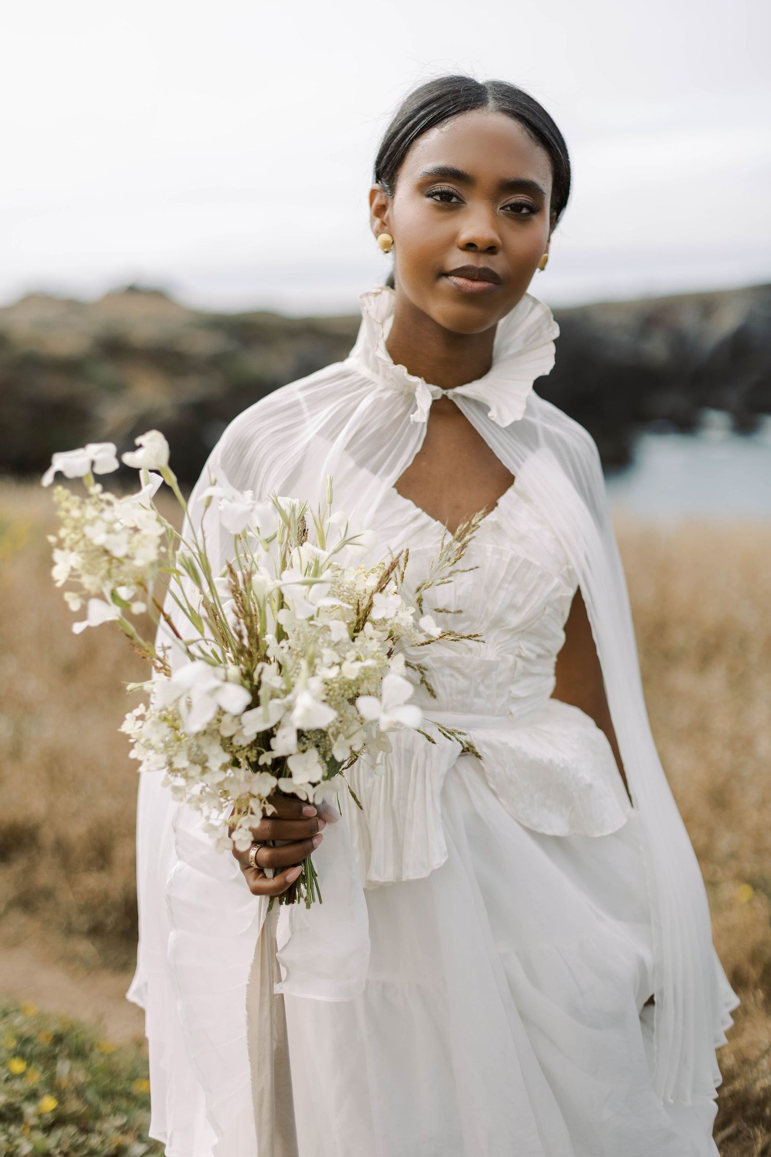 A bride in a flowing white dress holds delicate white flowers against a coastal landscape backdrop.