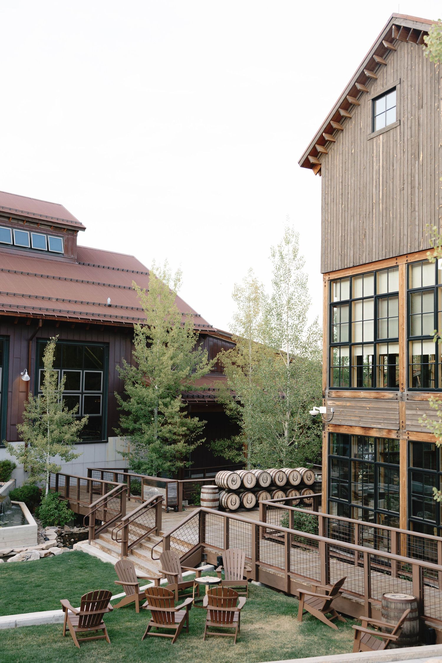 Modern rustic distillery building exterior with wooden deck and barrel decor against mountain backdrop.